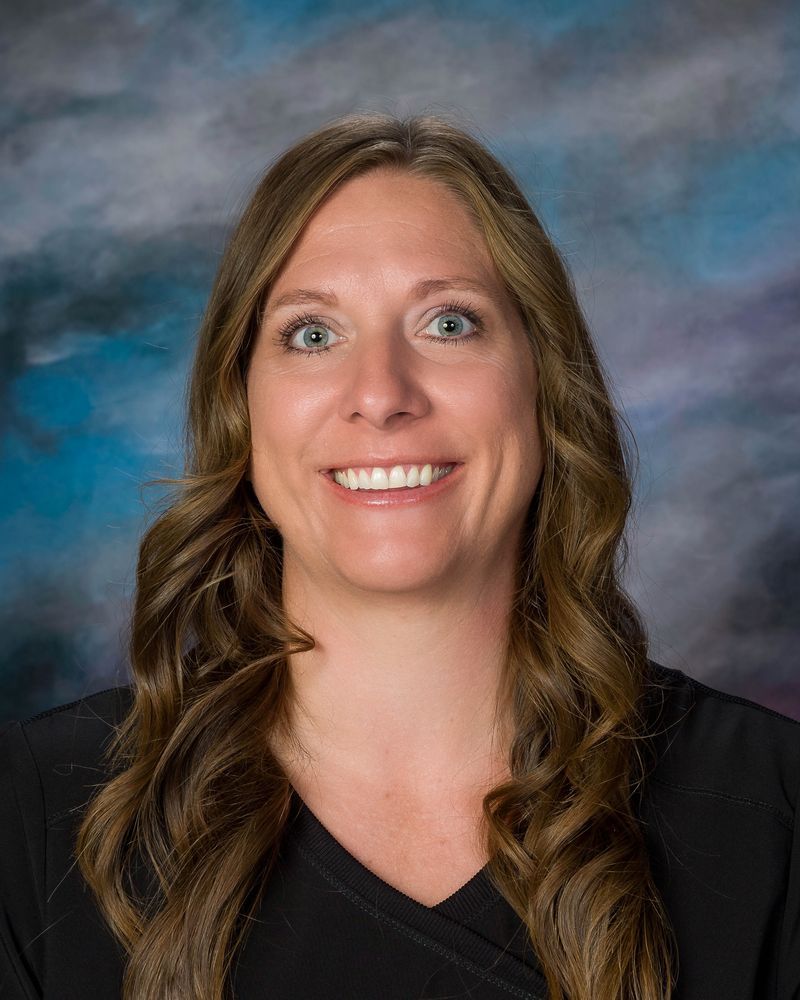 Smiling woman with long wavy brown hair, wearing a black shirt, in front of a blue and purple background.