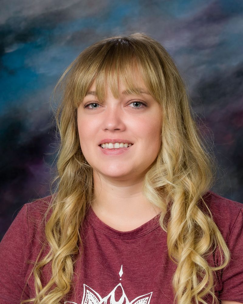 Woman with long blonde hair smiles, wearing a burgundy shirt against a blue and gray background.