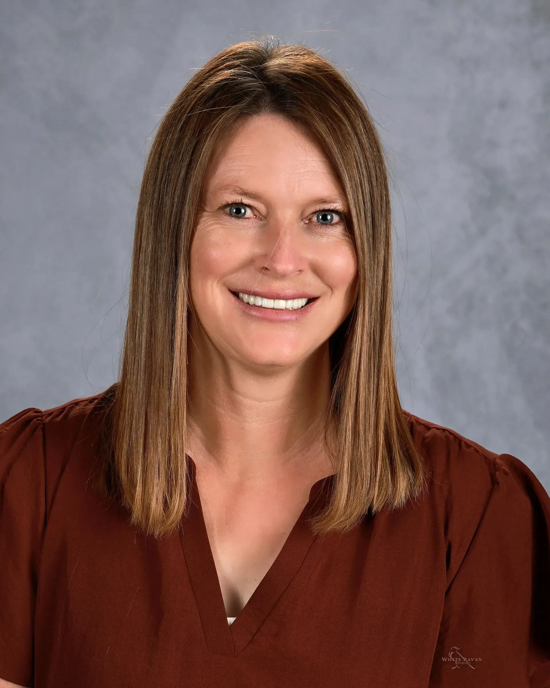 Woman with shoulder-length brown hair smiling, wearing a dark red top, set against a blurred gray background.