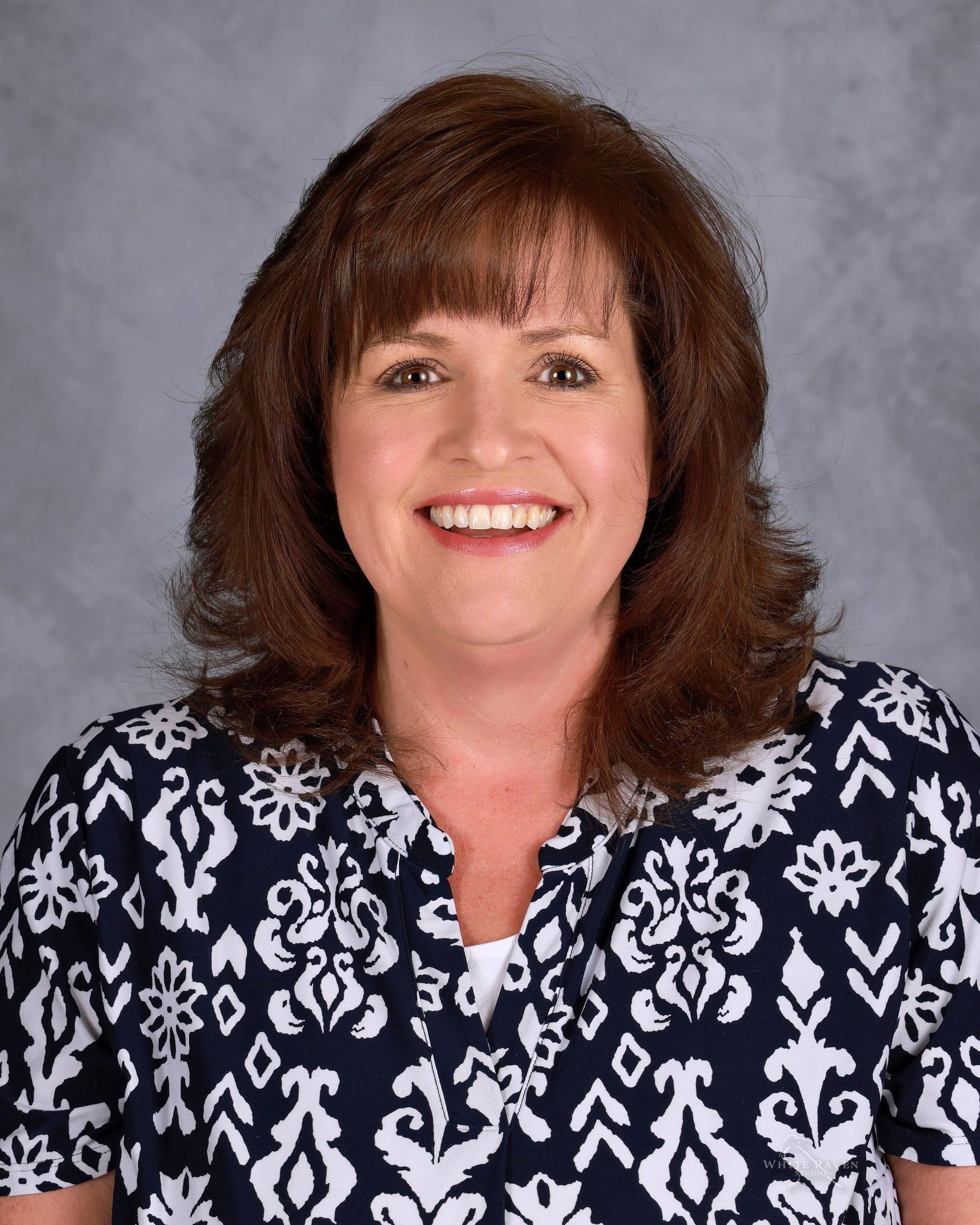 Woman with brown hair and bangs smiles, wearing a patterned blue and white blouse, against a gray background.