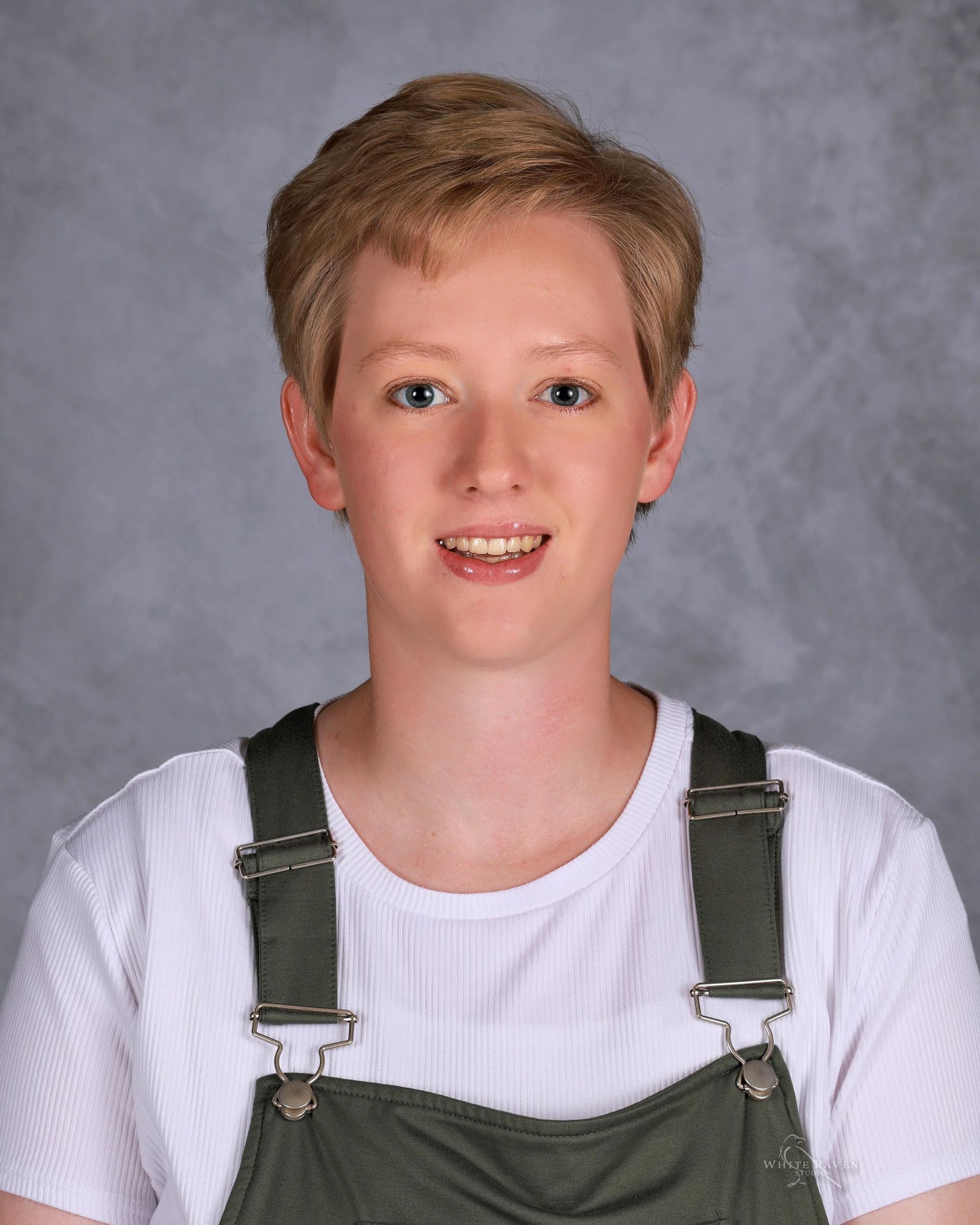 Woman with short blonde hair and a white shirt wearing green overalls, smiling.