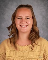 Smiling person with curly hair wearing a yellow top against a gray studio backdrop