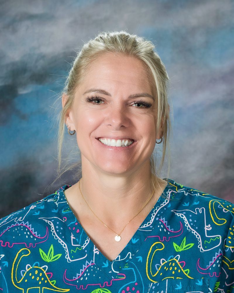 Woman smiling, wearing a turquoise dinosaur-print scrub top, small gold necklace, and hoop earrings.