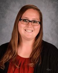 Studio portrait of a smiling woman with glasses, long brown hair, red top, and black cardigan on a gray background