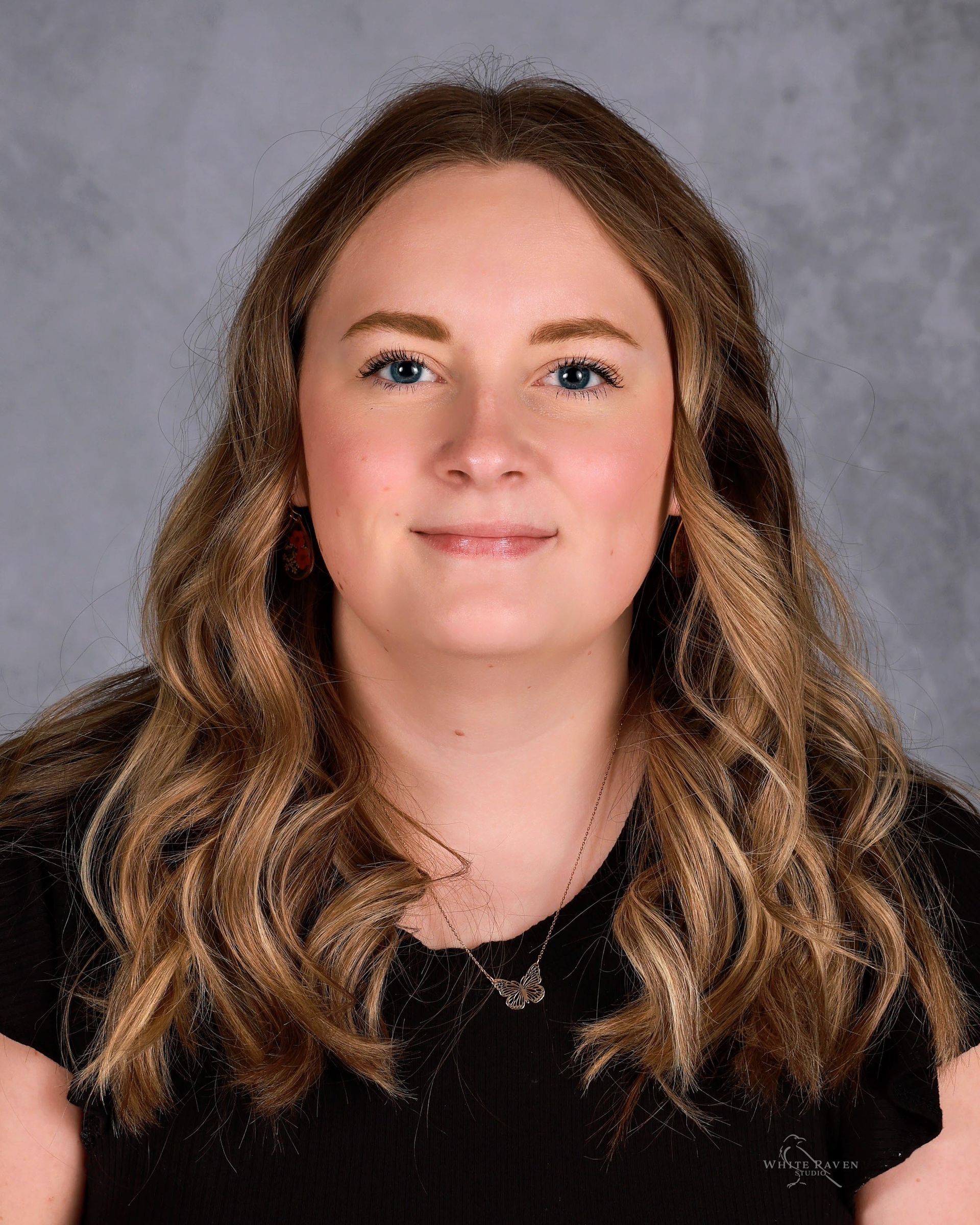 Woman with wavy blonde hair smiling, wearing a black shirt, posing in front of a gray backdrop.