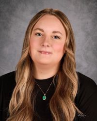 Portrait of a person with long light-brown hair, wearing a black top and necklace against a gray backdrop