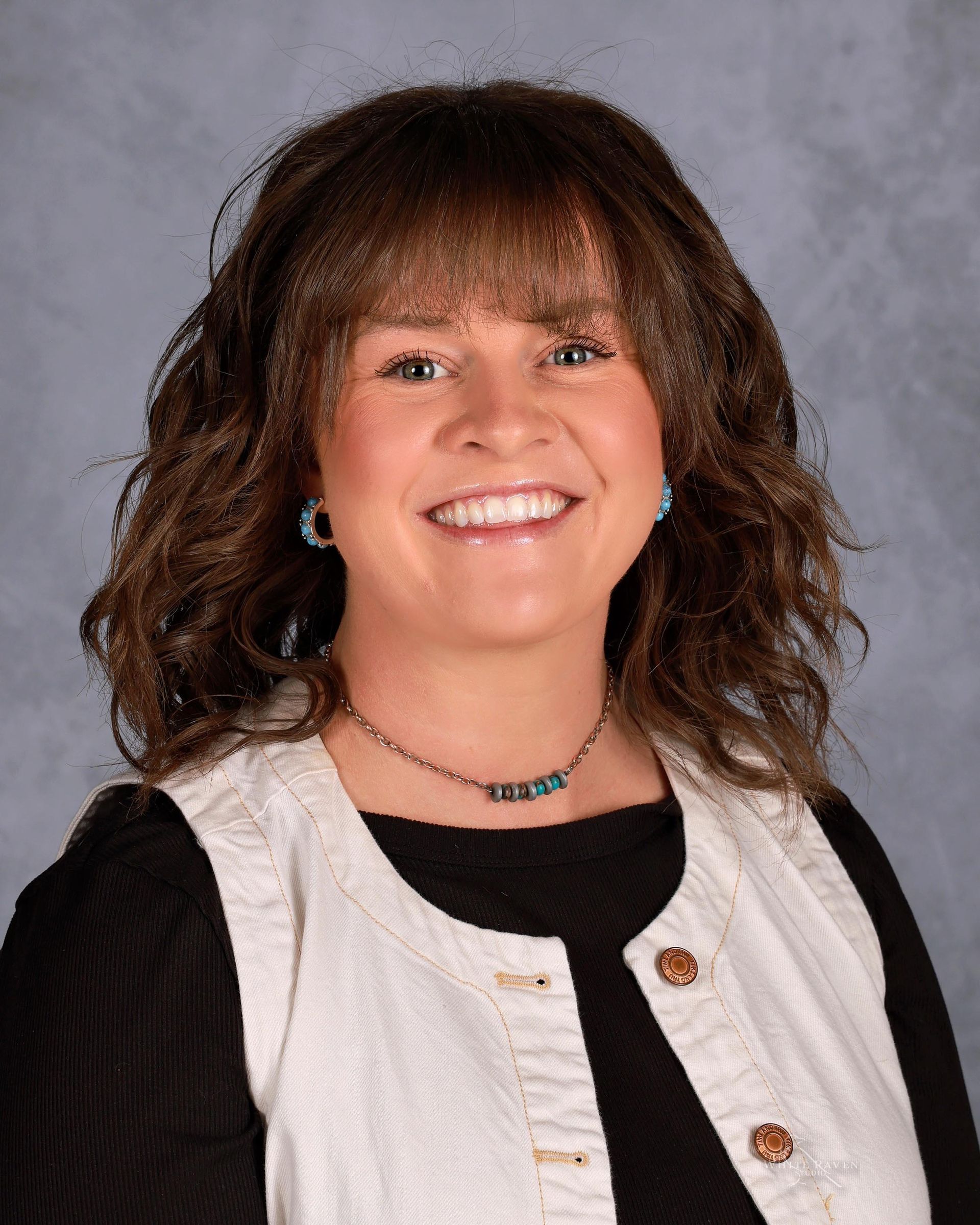 Woman with brown hair and bangs smiles, wearing a white vest over a black top, and jewelry.