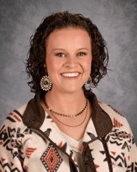 Smiling woman with curly hair wearing a patterned jacket against a gray studio backdrop