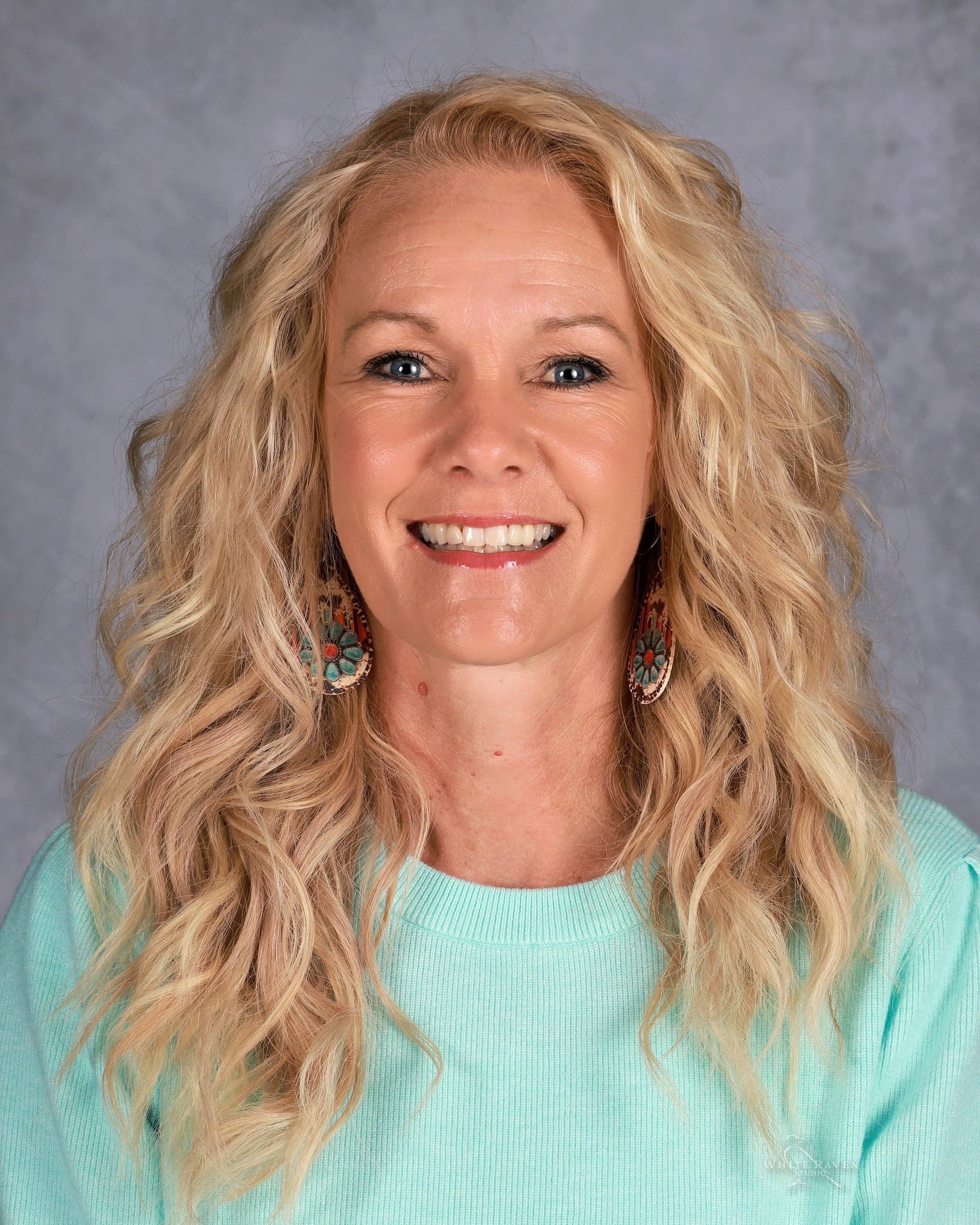 Woman with long blonde curly hair, smiling, wearing turquoise sweater, and earrings.