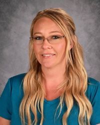 Smiling woman with long blonde hair and glasses, wearing a teal top against a gray studio backdrop