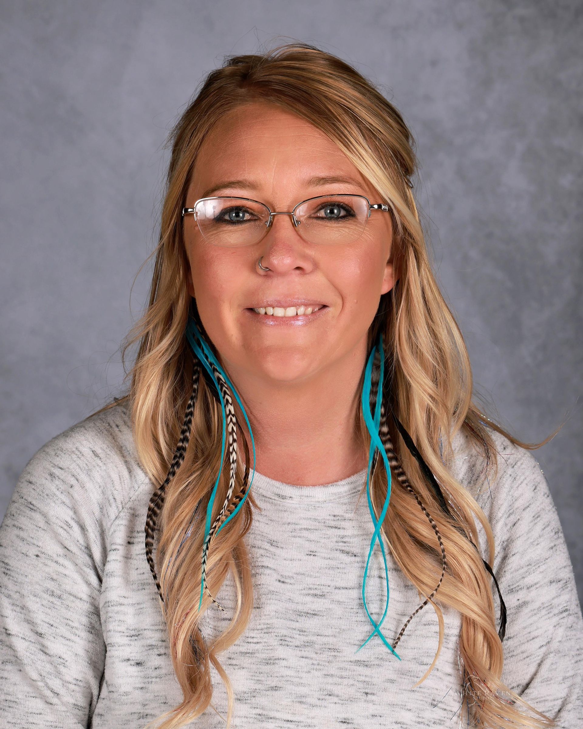 Woman with blonde hair, glasses, and blue feather hair extensions smiling in front of a gray backdrop.