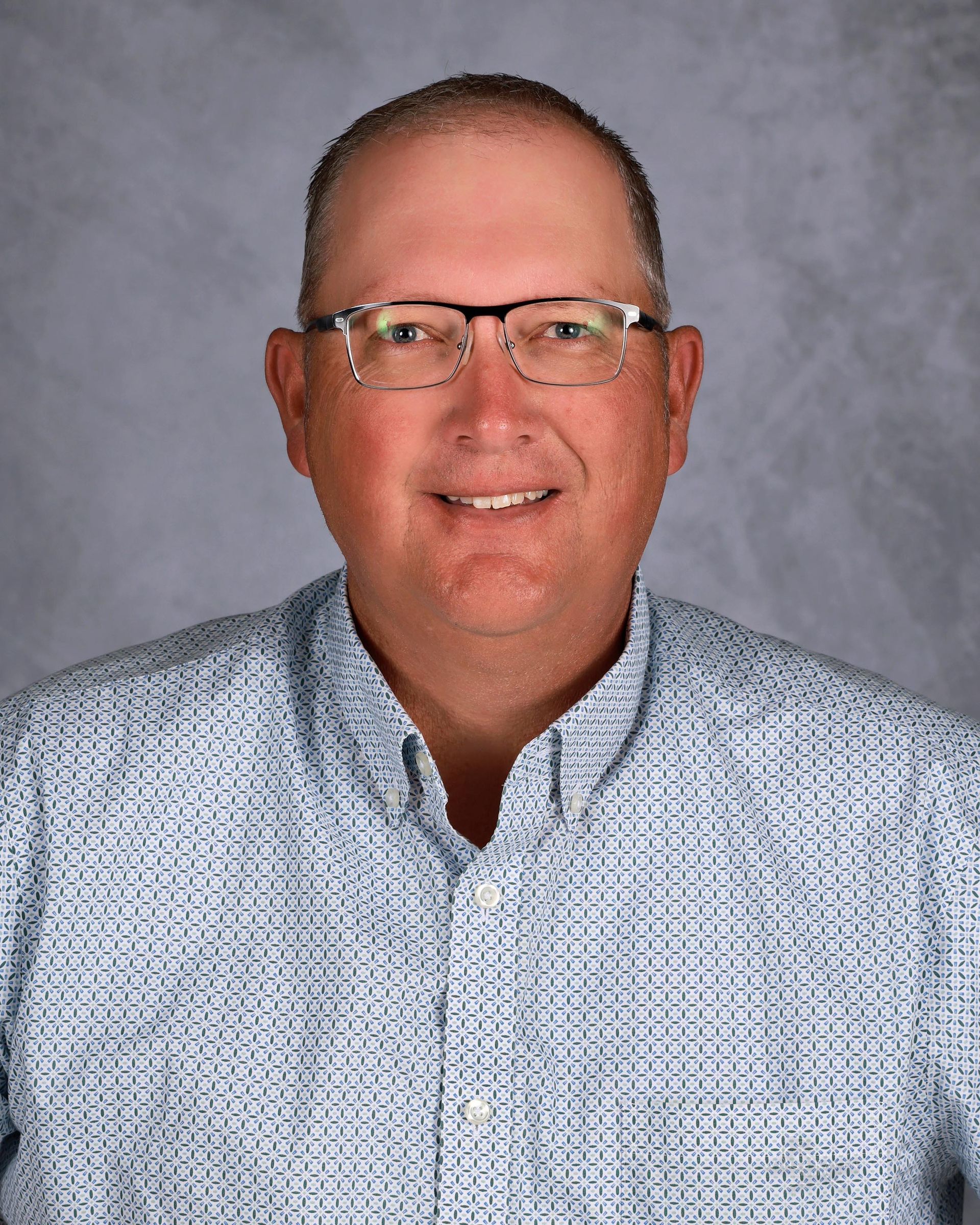 Man wearing glasses, smiling, in a patterned button-down shirt; studio portrait.