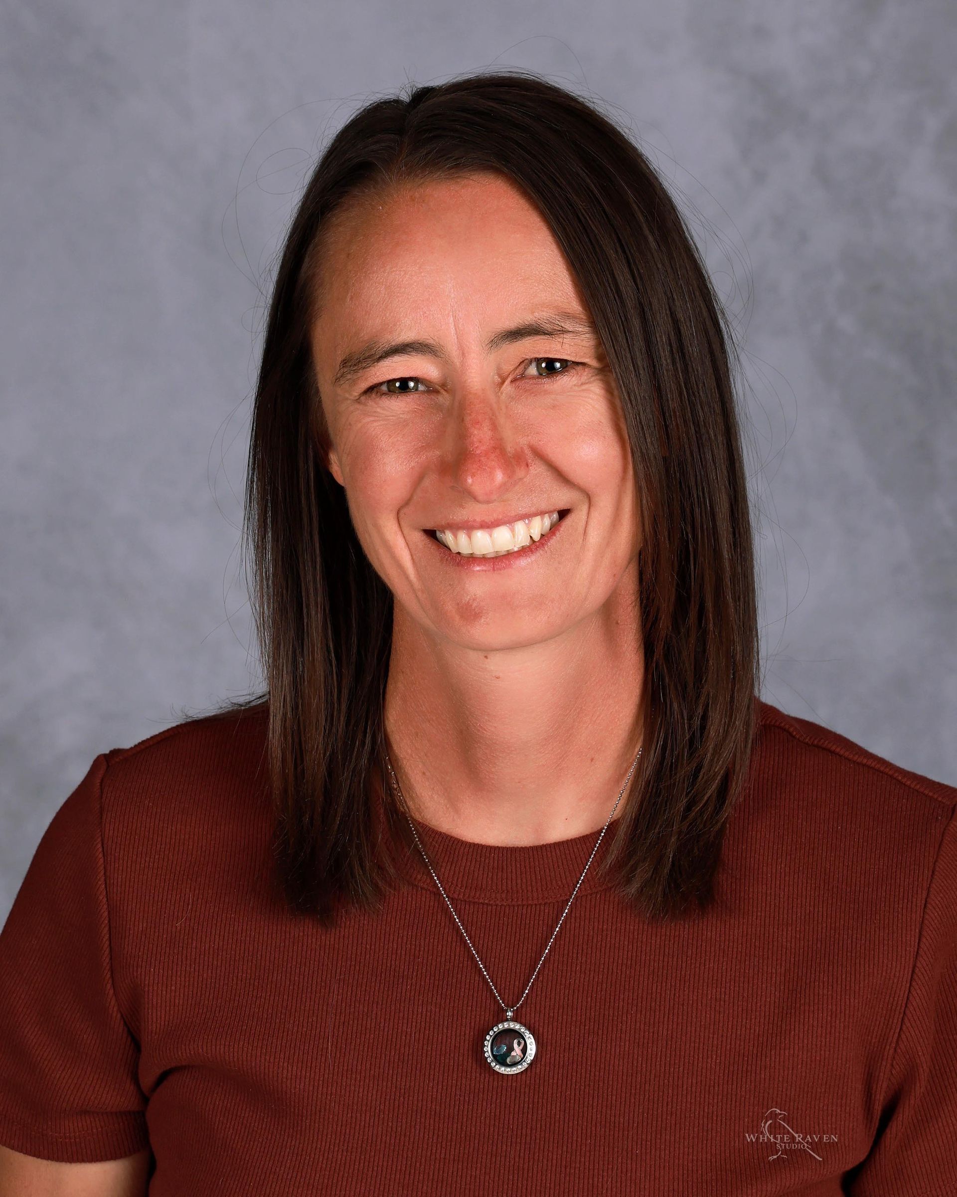Woman with brown hair, smiling, wearing a rust-colored shirt and a pendant necklace.