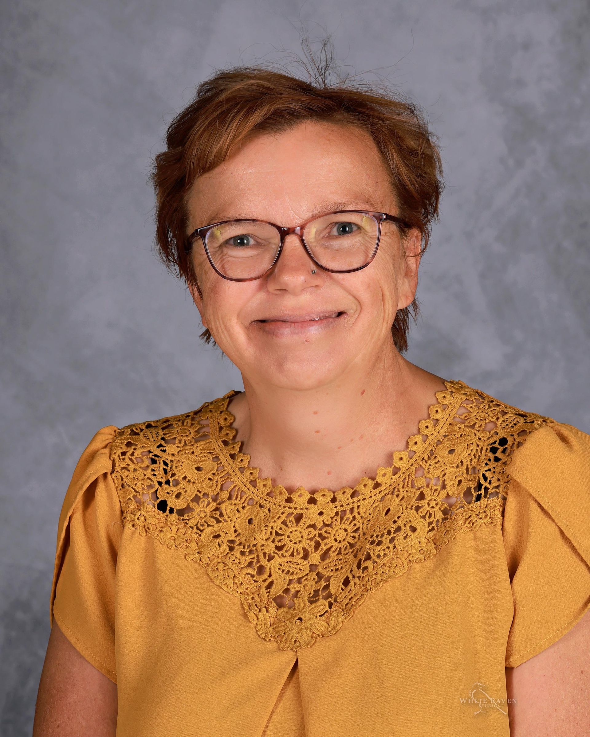 Woman wearing glasses and a yellow lace-collared top, smiling, against a mottled gray backdrop.