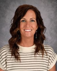 Smiling woman with wavy brown hair in a striped sweater against a gray studio backdrop