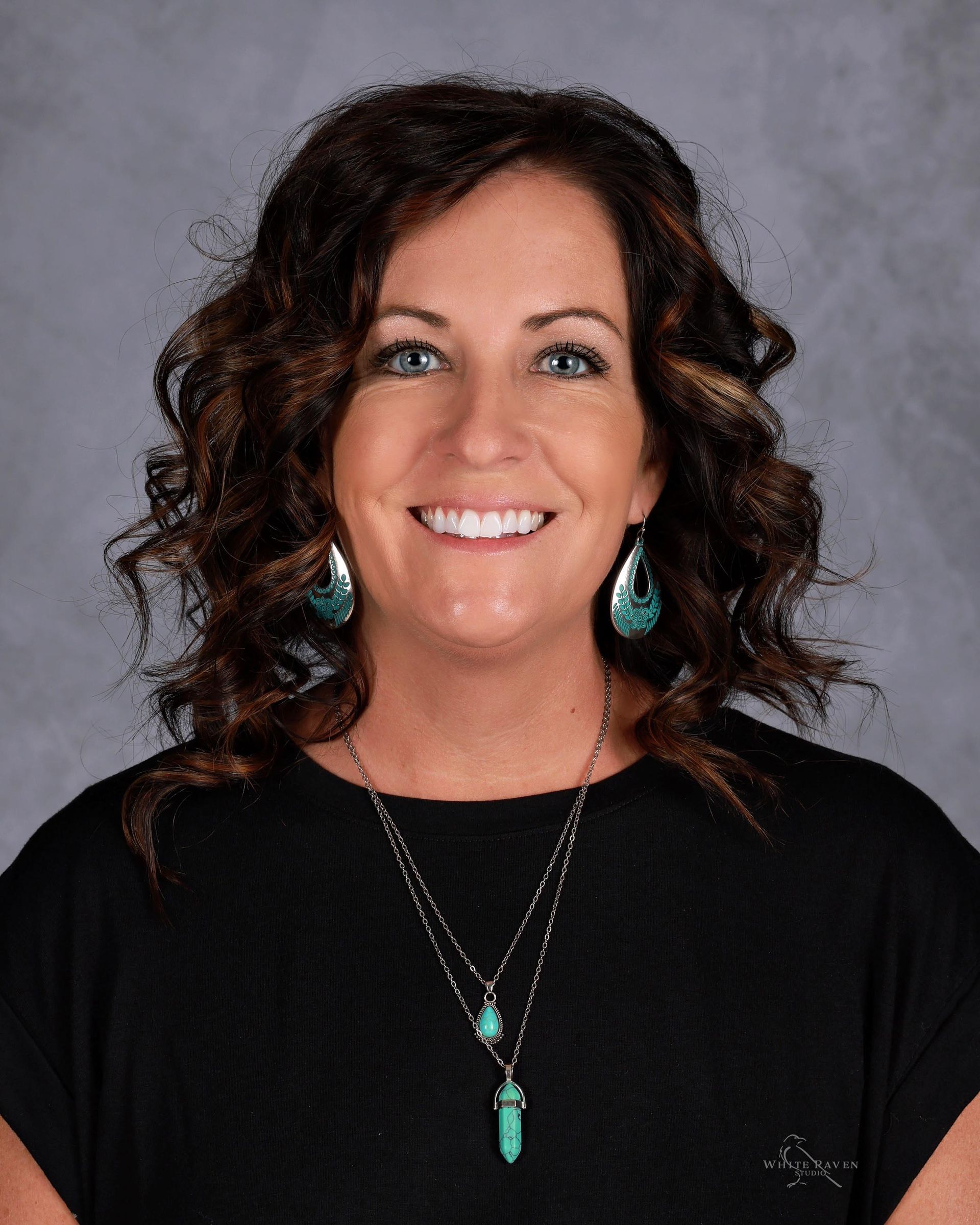 Woman with curly brown hair, wearing a black shirt, smiling, turquoise jewelry, and a gray background.