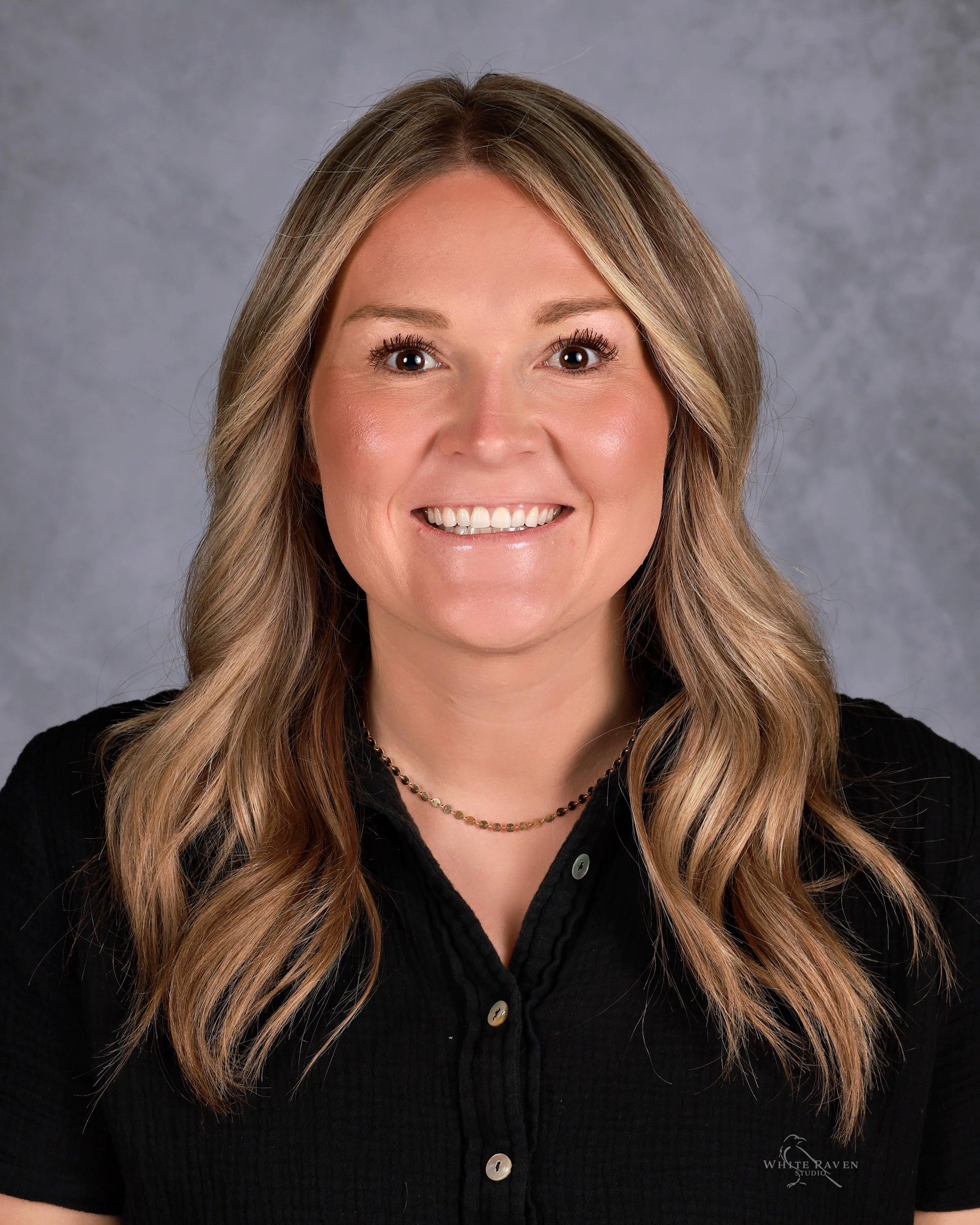 Woman with long blonde hair smiles, wearing a black shirt and a gold necklace, against a neutral backdrop.