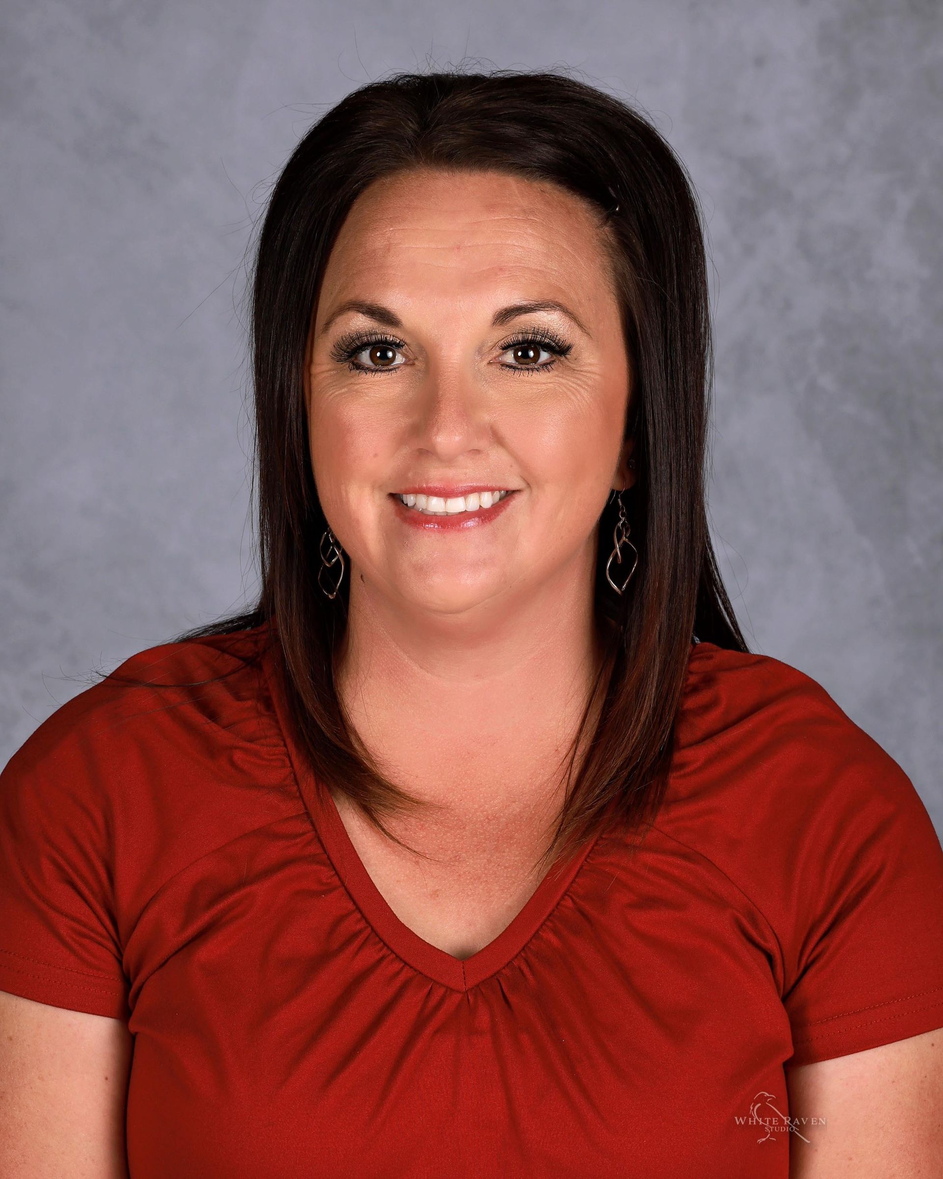 Woman with dark hair, wearing a red V-neck shirt, smiles at the camera against a gray background.