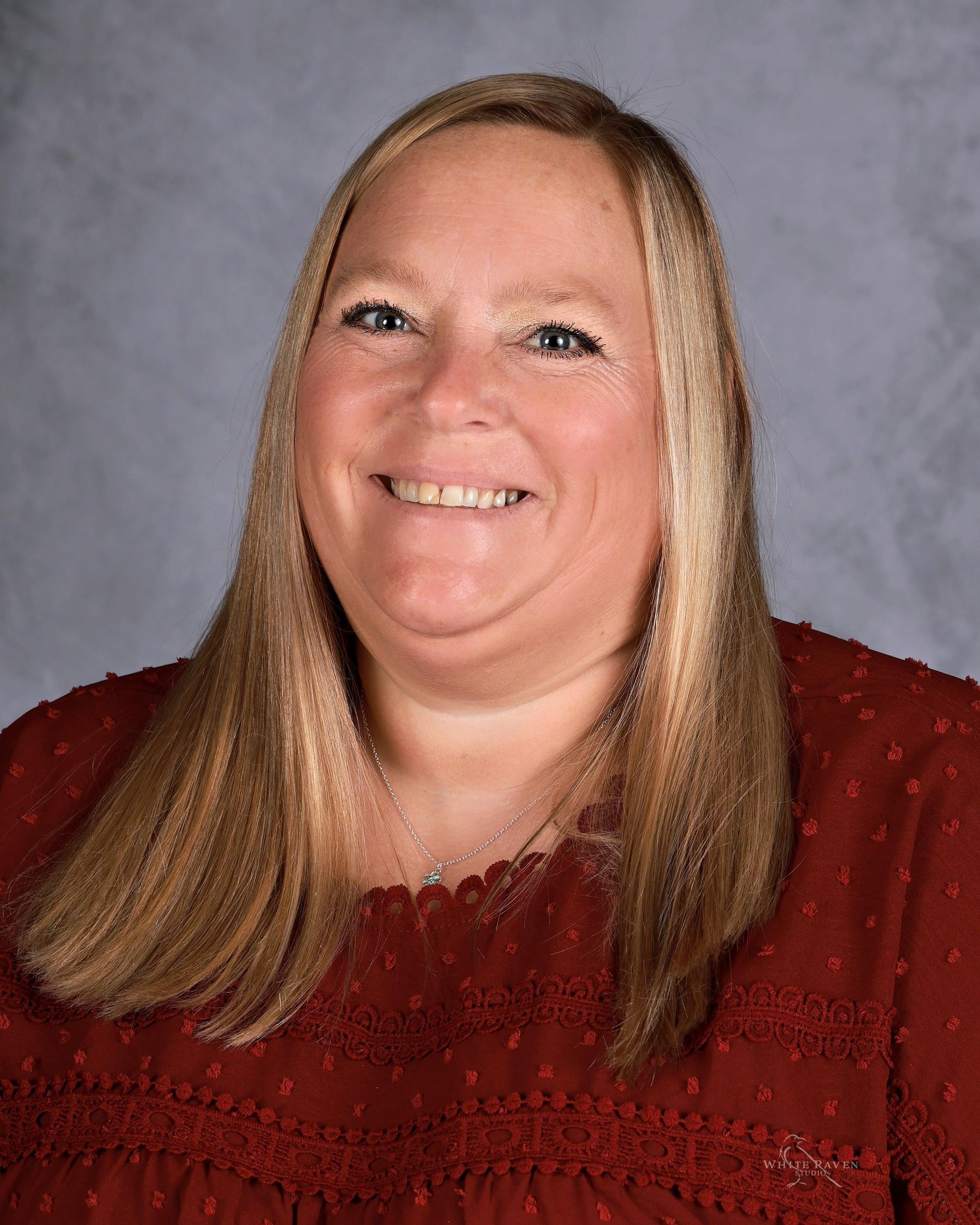 Woman with long blonde hair, smiling, wearing a red top, against a gray background.
