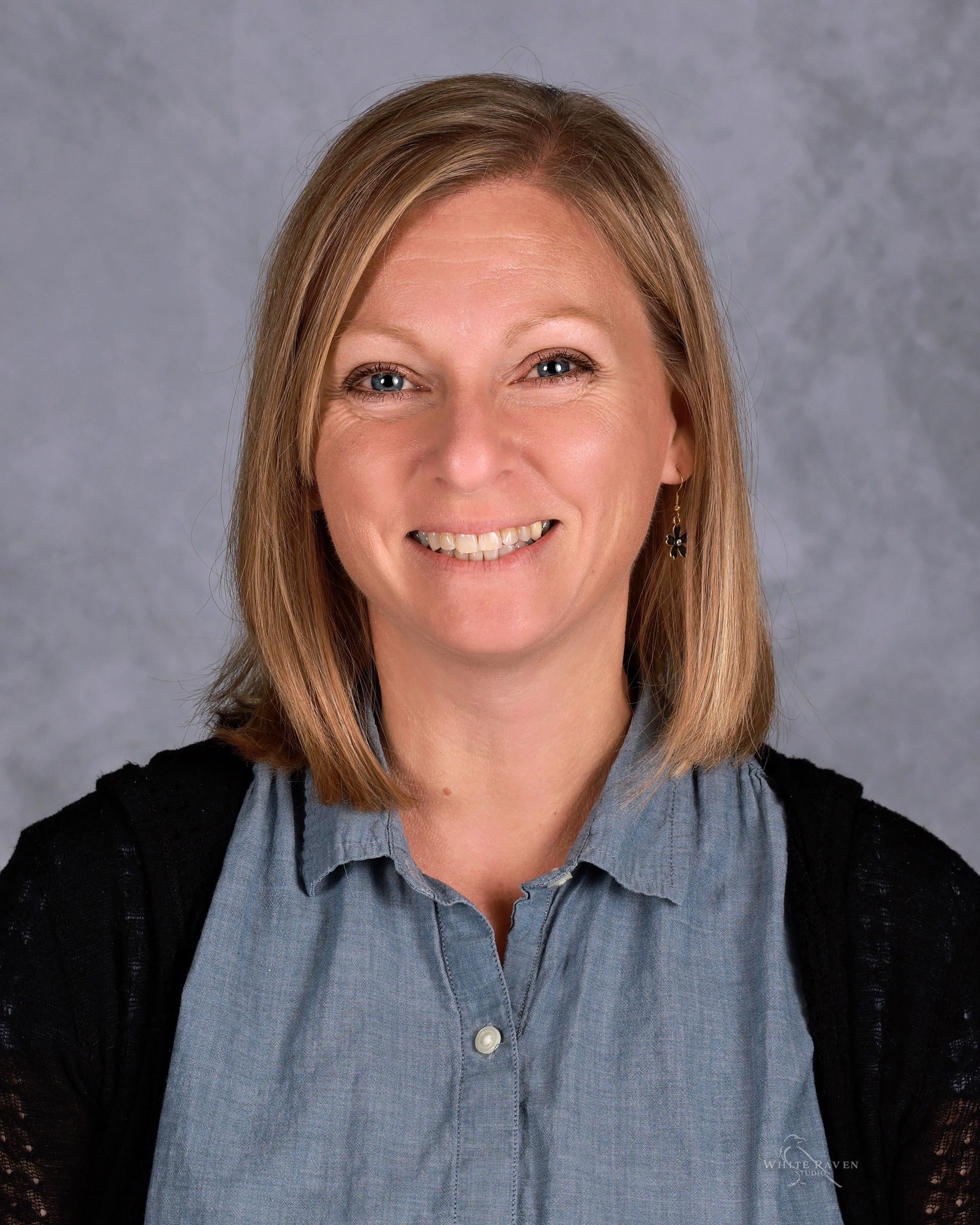 Woman with blonde hair, smiling, wearing a denim shirt and black cardigan, against a gray backdrop.