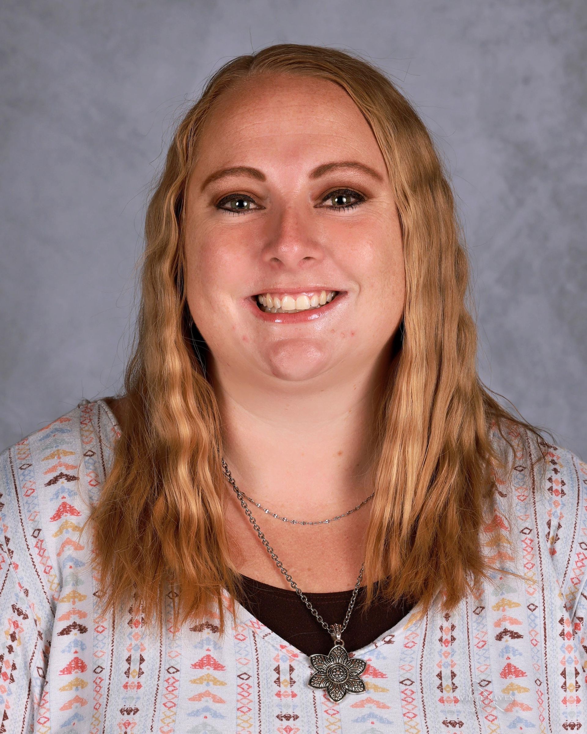 Woman with long blonde hair, smiling, wearing a patterned top and necklace, set against a blurred background.