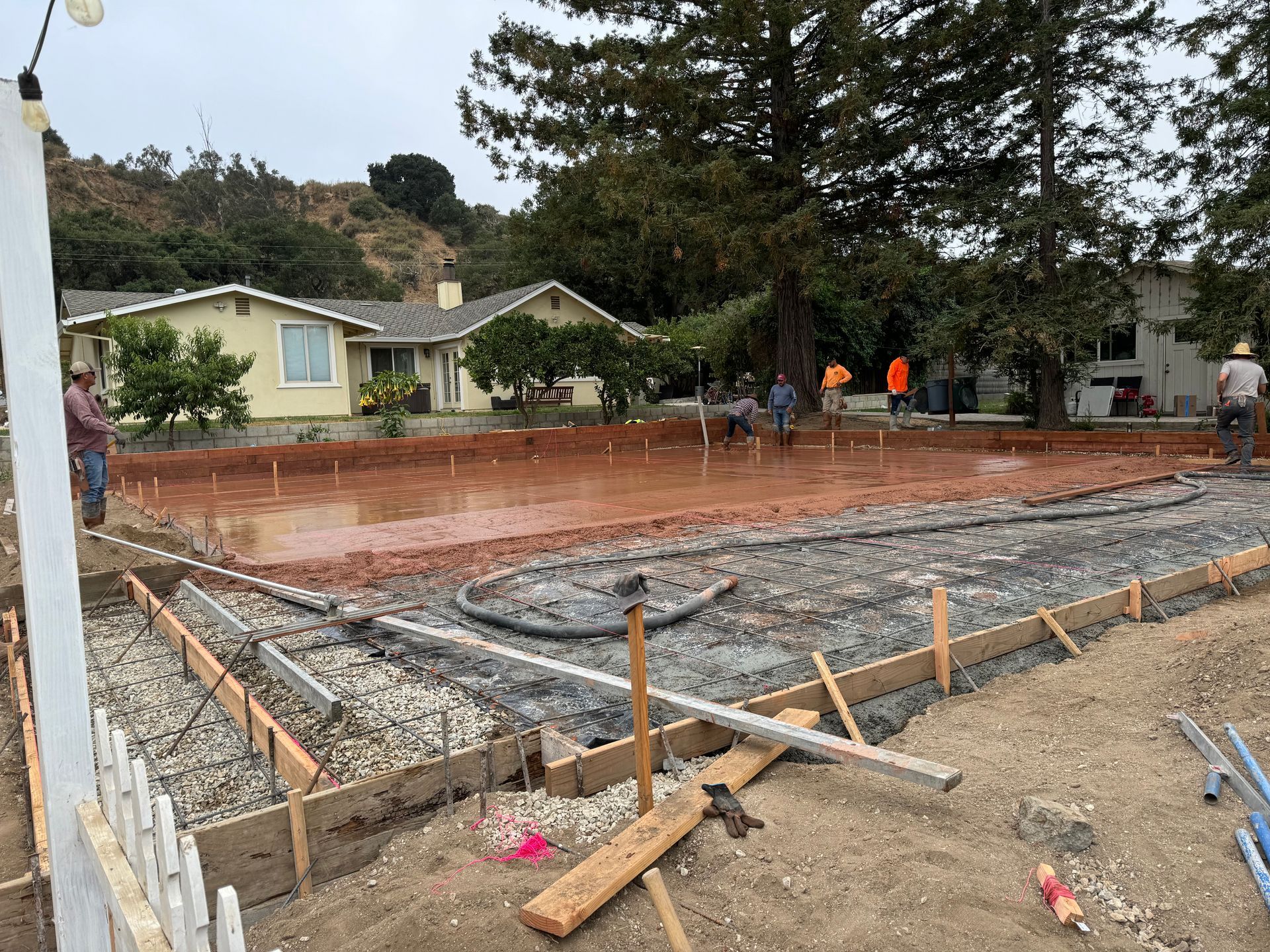 Concrete foundation under construction beside a suburban house, with workers and wooden forms around the site.