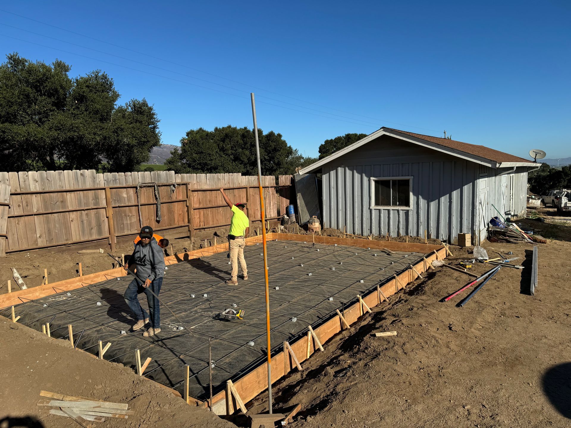 Workers pour a concrete foundation in a fenced backyard beside a small shed.