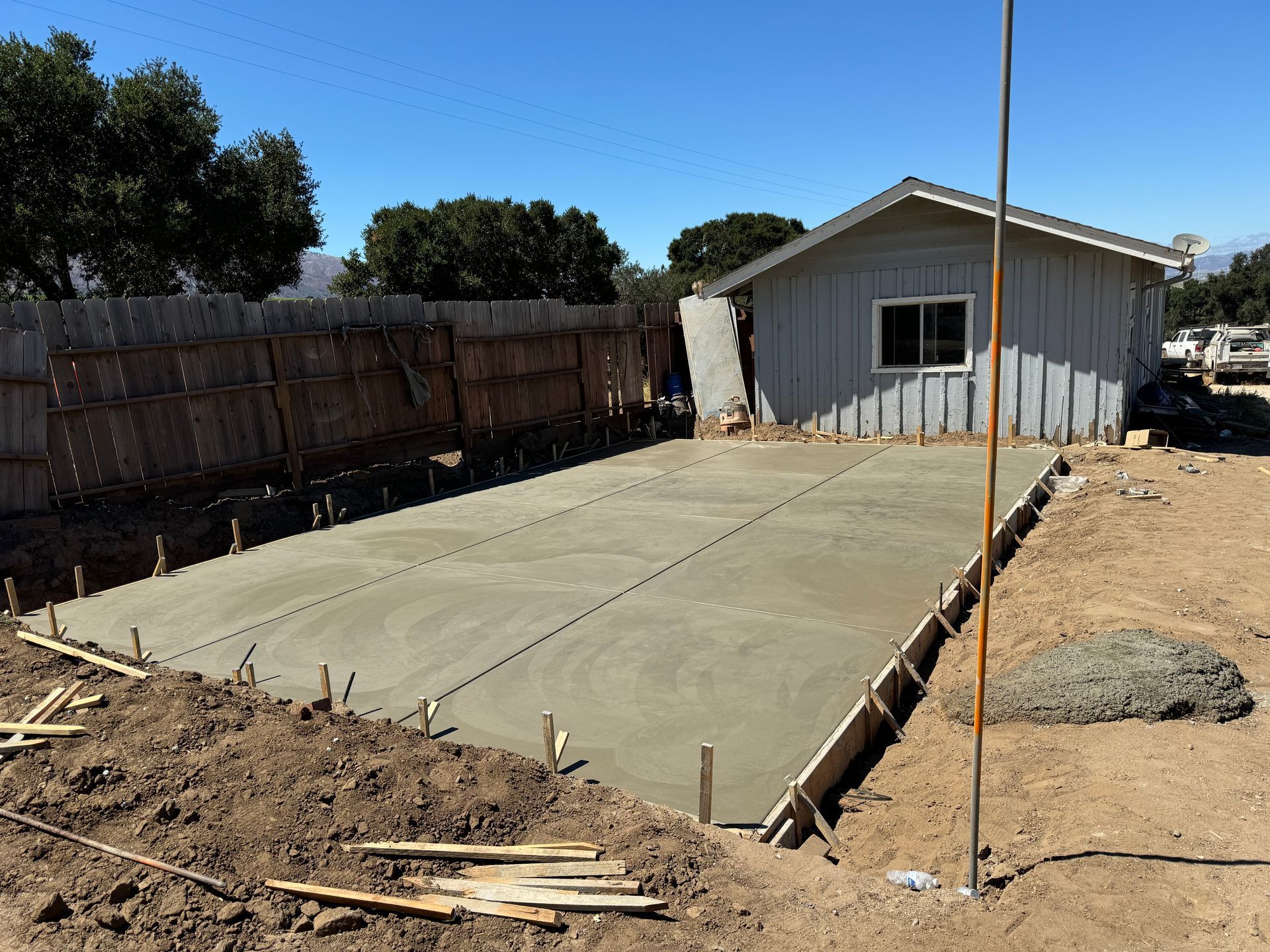 Concrete slab patio area beside a gray house under construction in a dirt yard, with wooden fencing and trees.