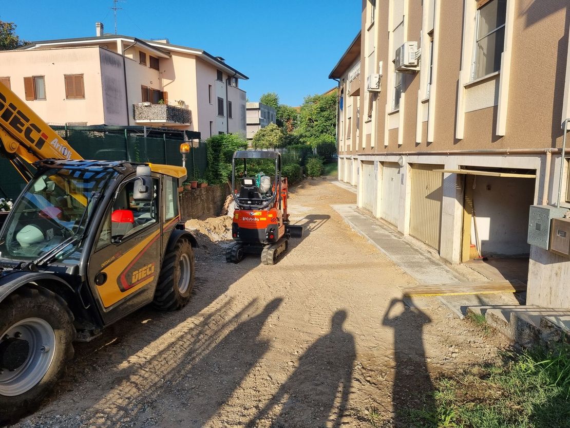 Cantiere: un carrello elevatore giallo e un escavatore arancione su un sentiero di ghiaia vicino a un edificio con garage.