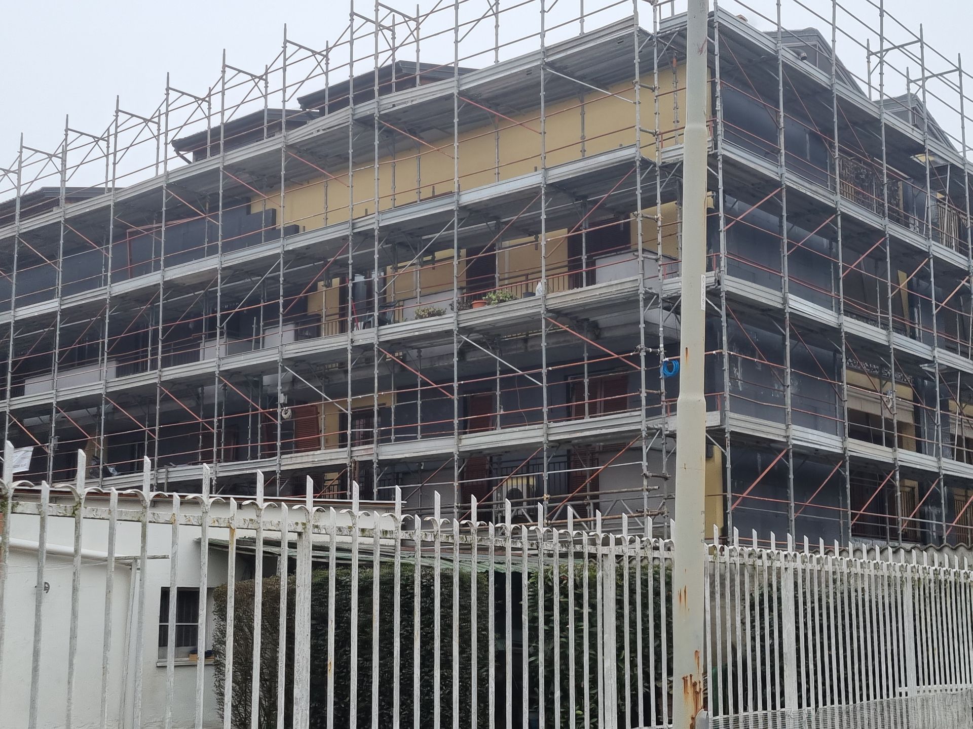 Edificio in ristrutturazione, coperto da impalcature, dietro una recinzione bianca.