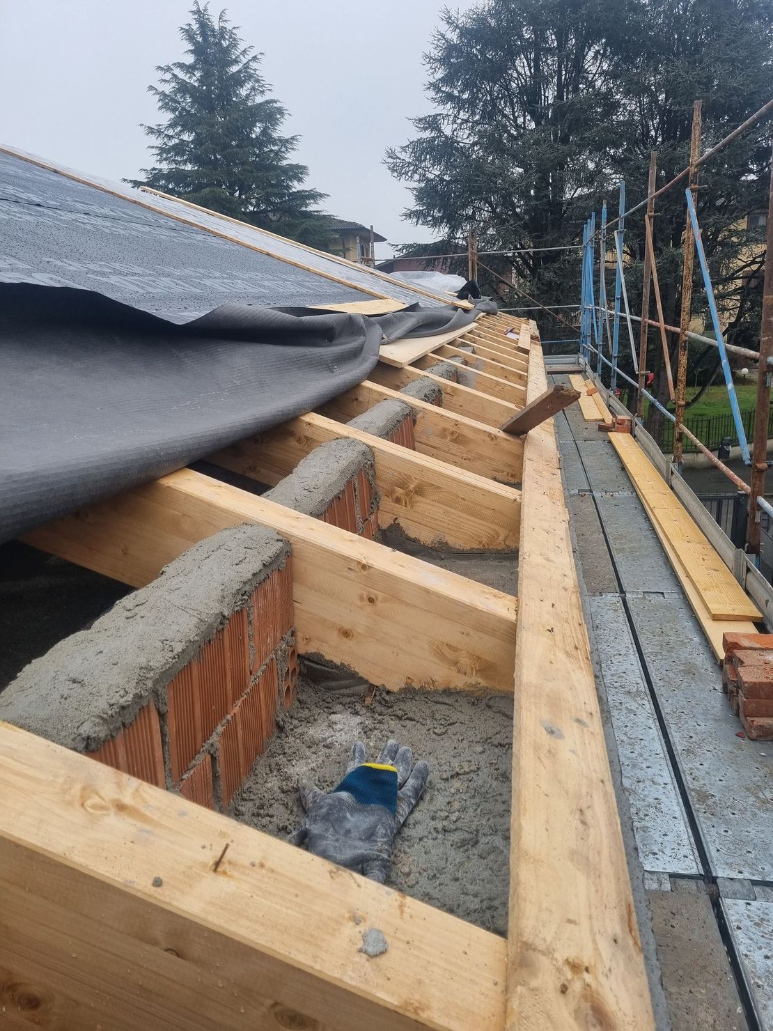 Costruzione del tetto in corso: travi in legno, mattoni e cemento grigio, con materiale di copertura nero parzialmente installato.