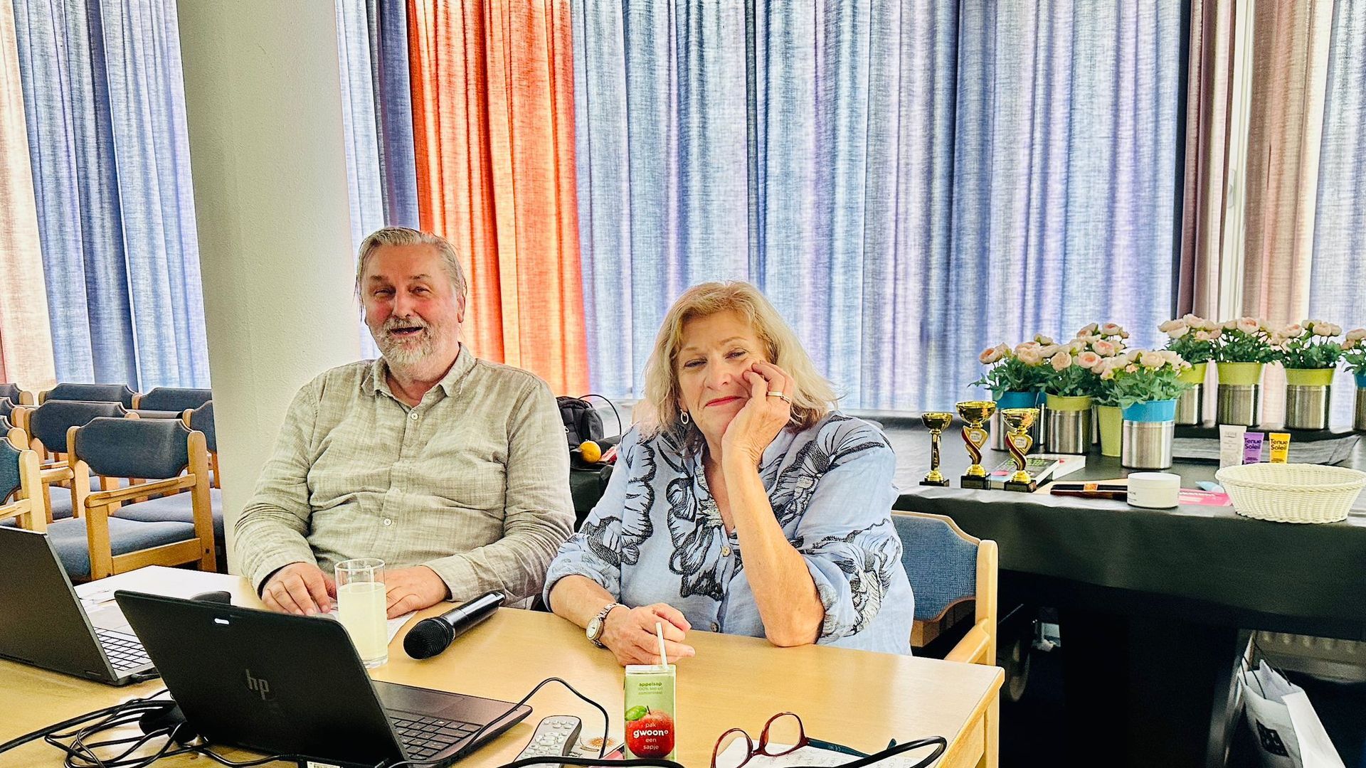 Twee mensen aan een tafel met laptops en microfoons. Vrouw in een blauwe top met bloemenprint. Man in een beige overhemd.
