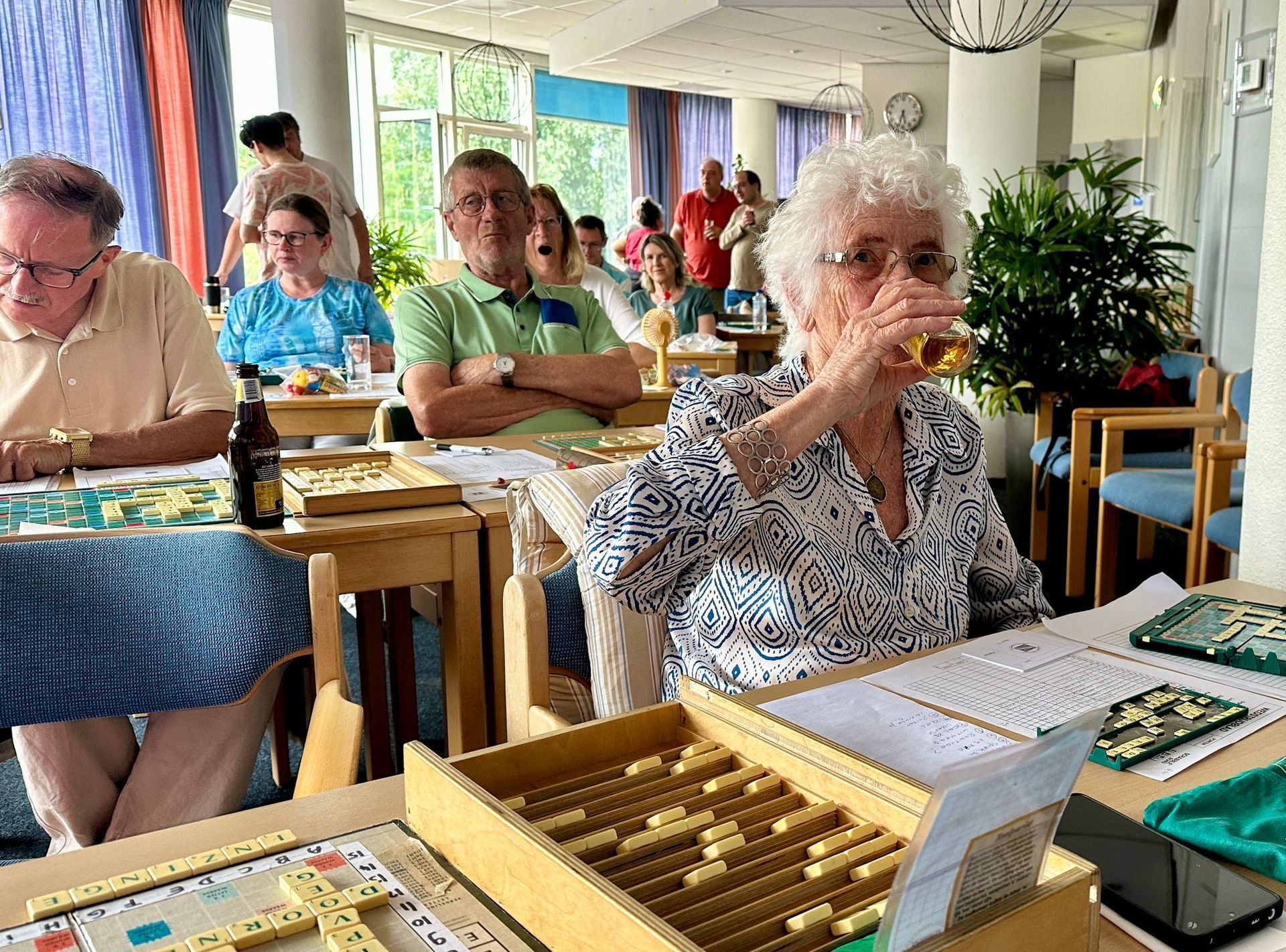 Mensen spelen bordspellen in een goed verlichte kamer; een vrouw drinkt uit een glas.