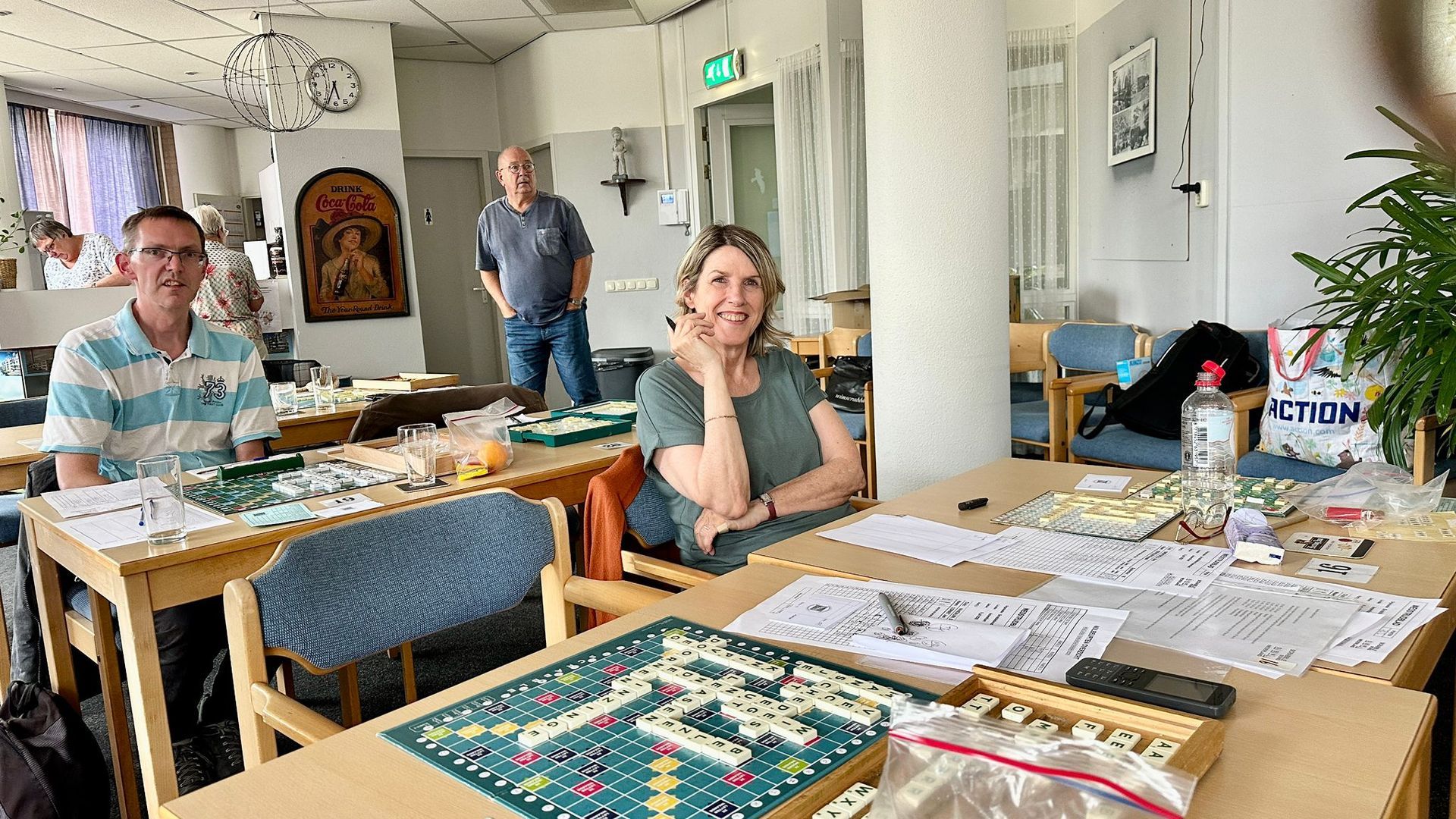 Drie mensen zitten aan tafels en spelen spelletjes in een felverlichte ruimte. Twee glimlachen, één kijkt toe.