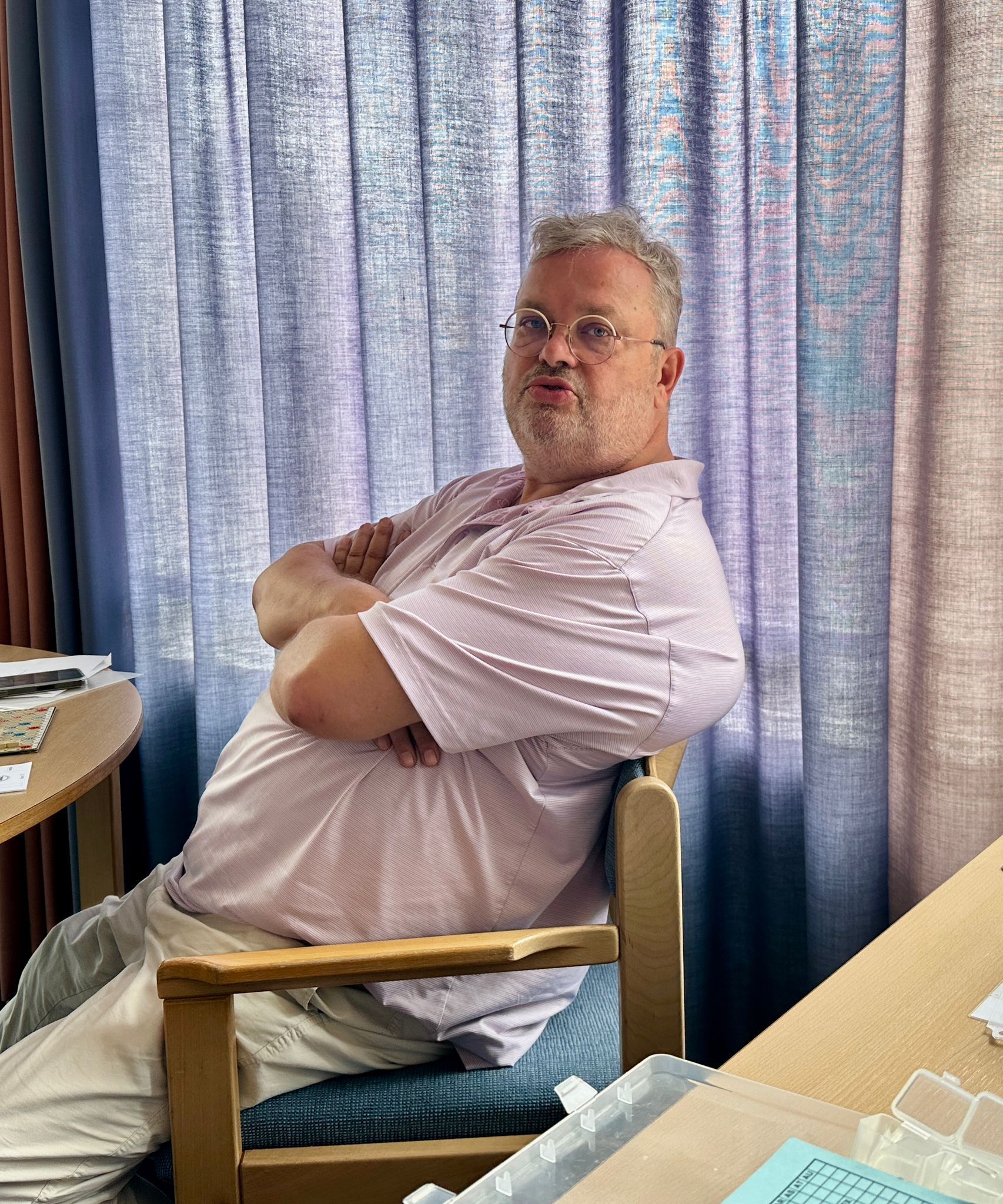 Een man met een bril zit met de armen over elkaar voor een gordijn. Hij draagt ​​een lichtroze shirt.