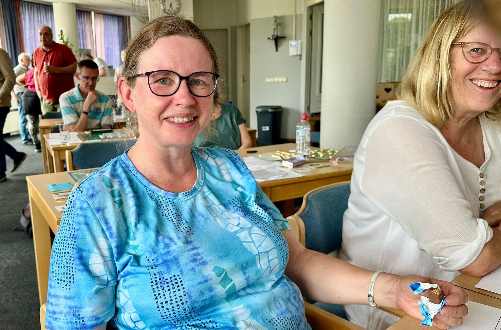 Een lachende vrouw, met een blauw tie-dye shirt en een bril, zit aan een tafel. Op de achtergrond zijn anderen te zien; het is een binnenruimte.
