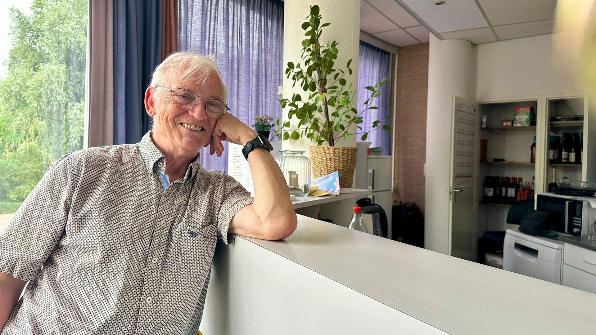 Een glimlachende man leunt tegen een toonbank, met zijn arm rustend, binnen. Achter hem is een raam en een plant te zien.
