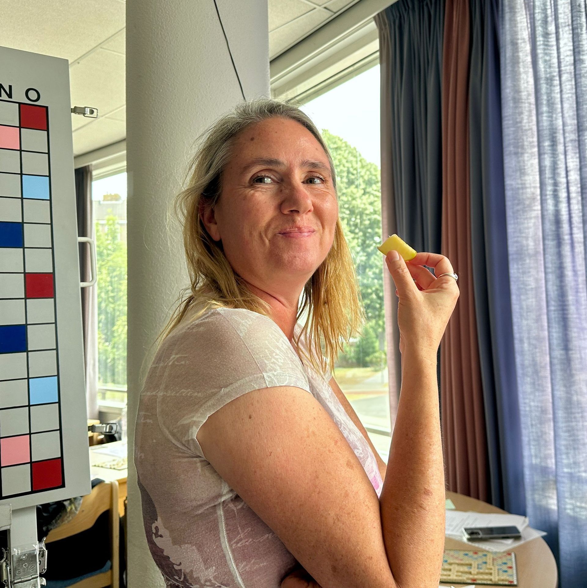 Een vrouw met een geel snoepje in haar hand staat lachend voor een raam. Links hangt een kleurrijk bord.