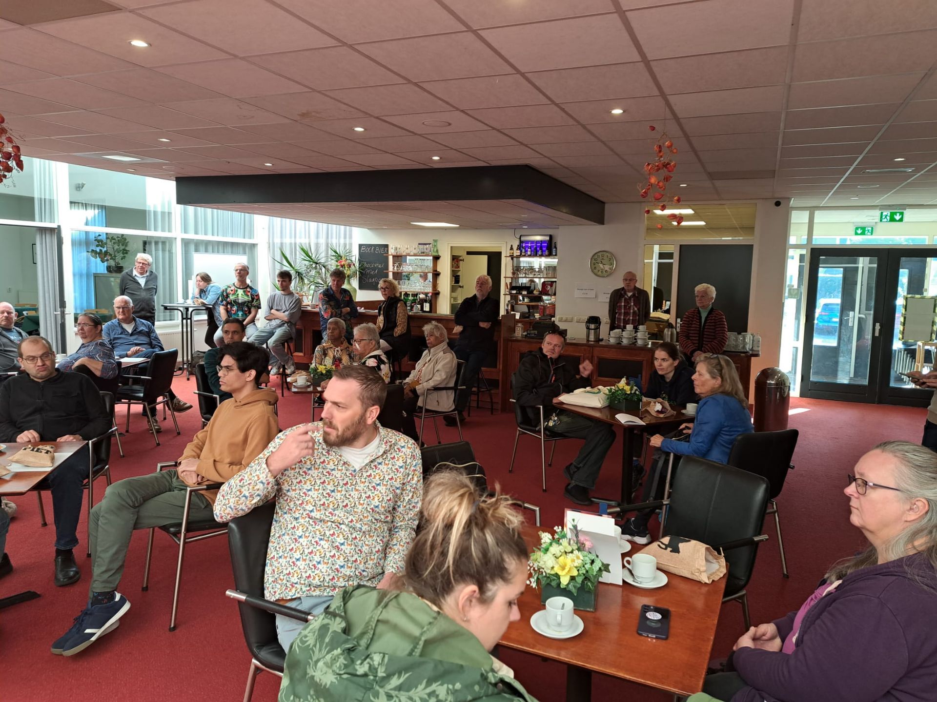 Een groep mensen zit aan tafels, waarschijnlijk tijdens een vergadering in een goed verlichte binnenruimte.