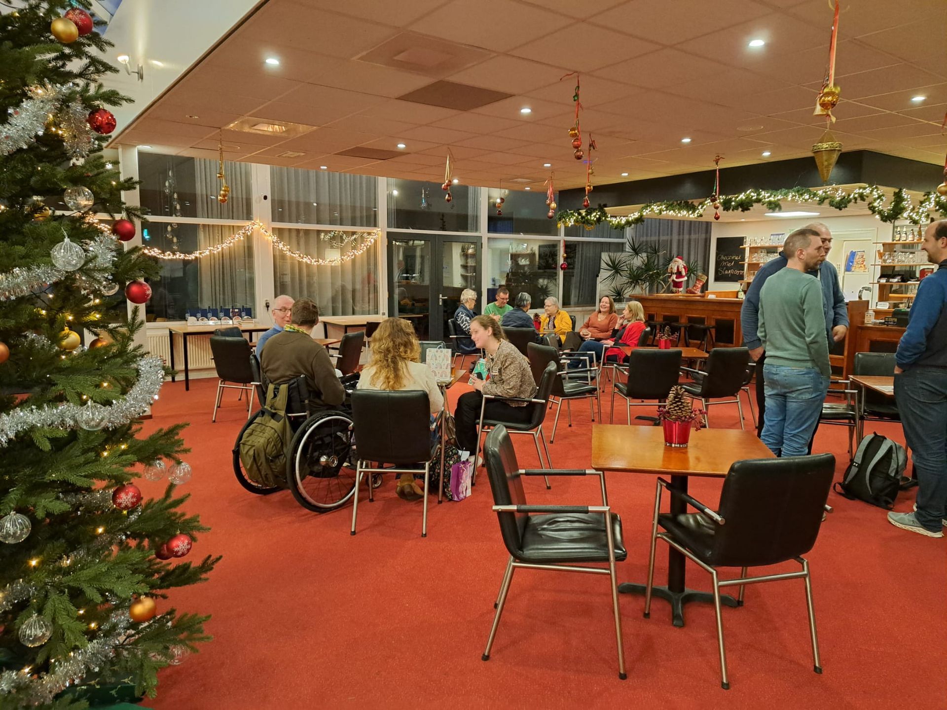 Interieur van een versierde kamer waar mensen met elkaar praten; sommigen zitten in een rolstoel. Er staat een kerstboom in de buurt.