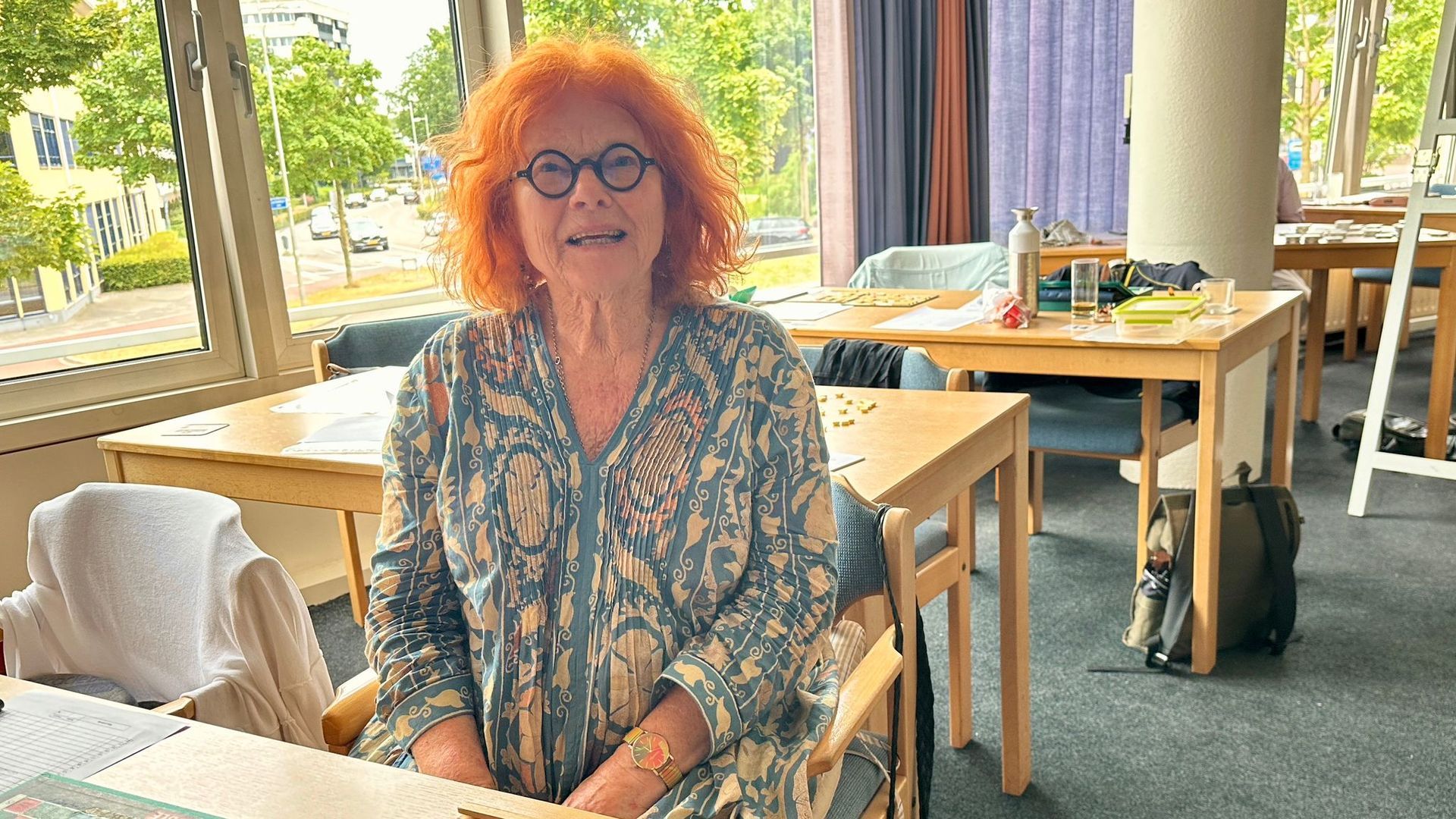 Een vrouw met rood krullend haar en een bril zit glimlachend aan een tafel in een lichte kamer.
