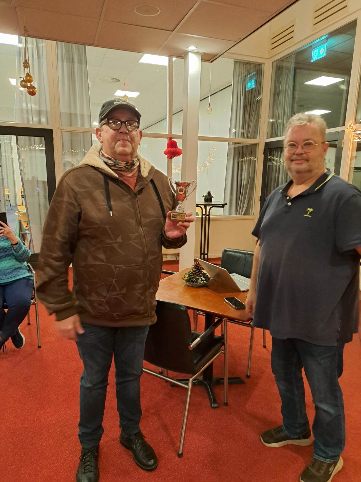 Twee mannen staan ​​binnen, een van hen houdt een trofee vast. Ze staan ​​vlak bij een tafel met een microfoon en een rode loper.
