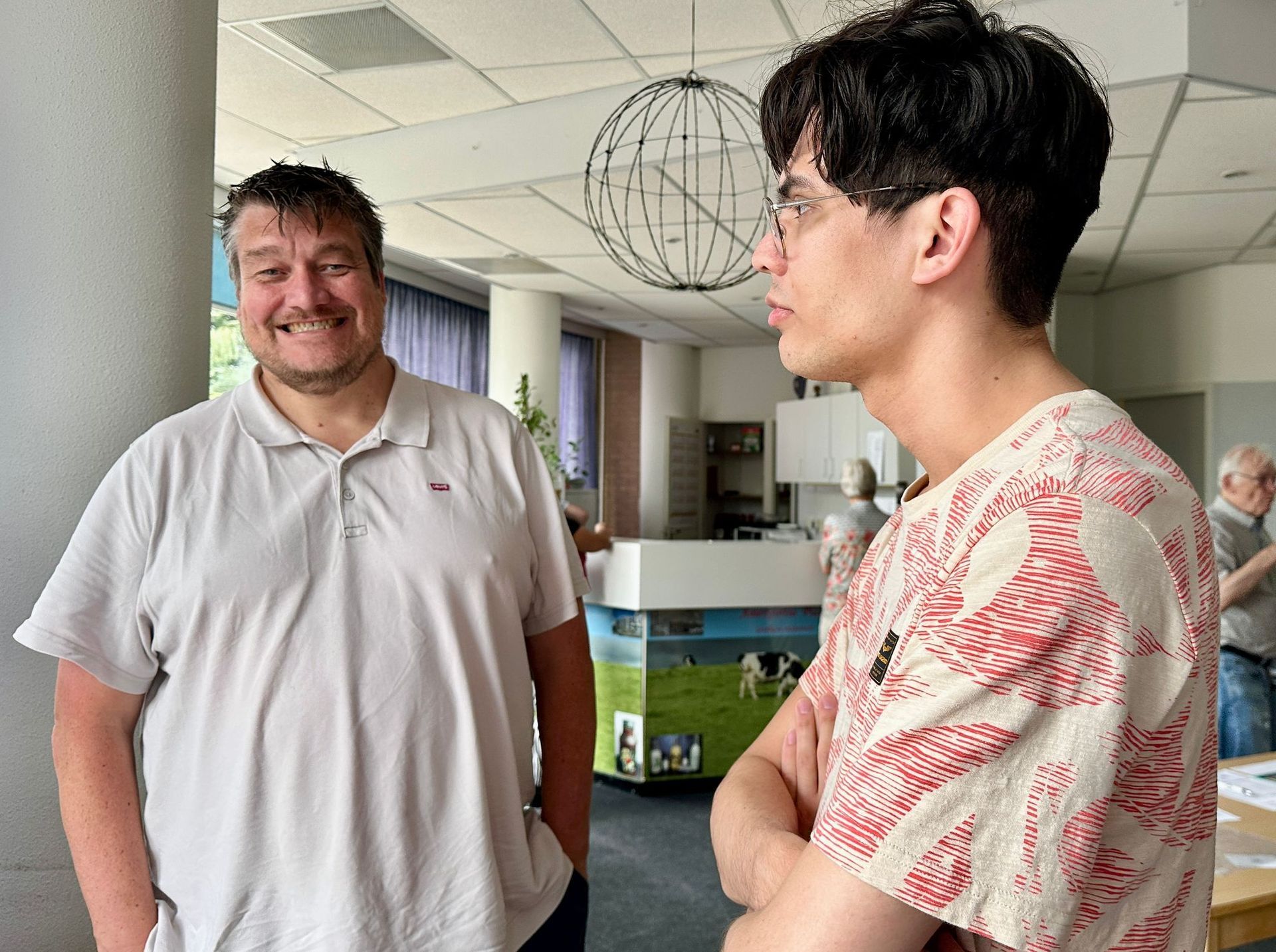 Twee mannen in een lichte kamer. De ene glimlacht, gekleed in een poloshirt. De andere praat, gekleed in een T-shirt met een patroon.