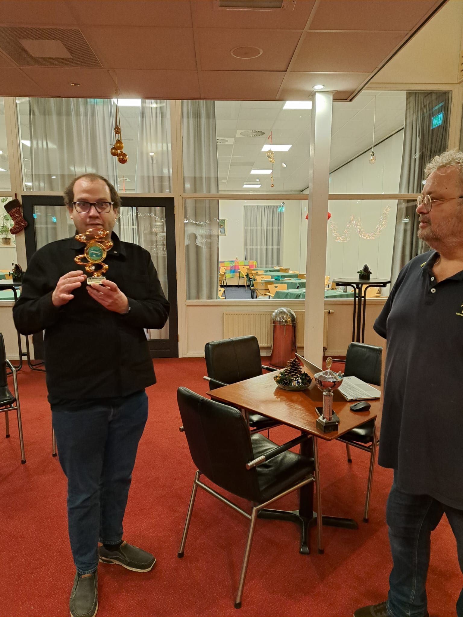 Een man in zwart met een trofee in zijn hand staat naast een andere man in een kamer met een tafel en stoelen.