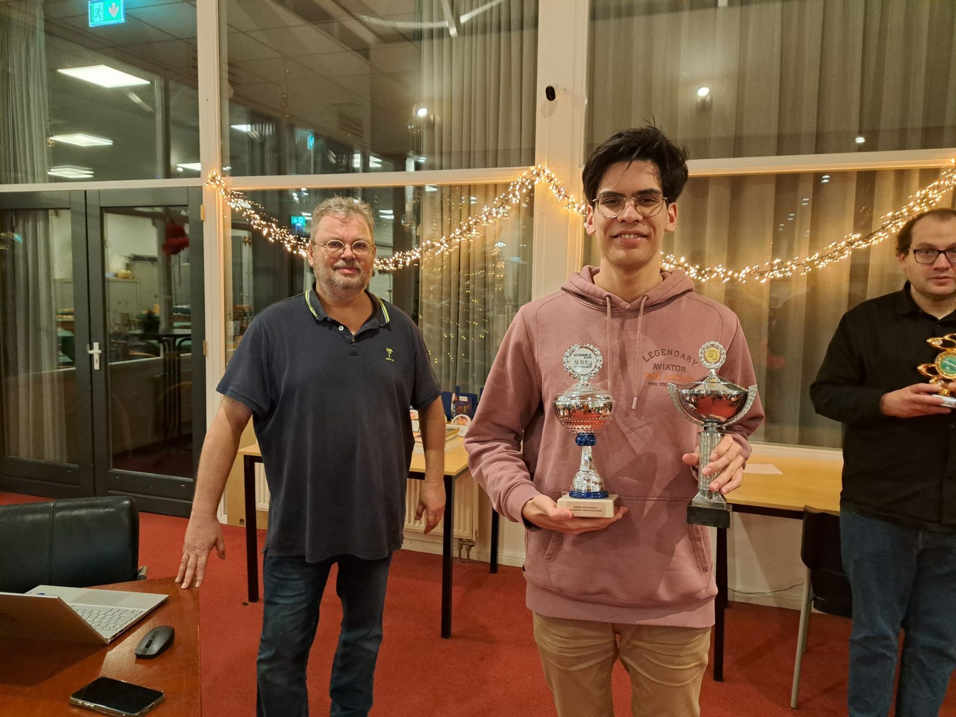 Drie mannen poseren binnen, een van hen houdt twee trofeeën vast, een ander een trofee, de anderen glimlachen.