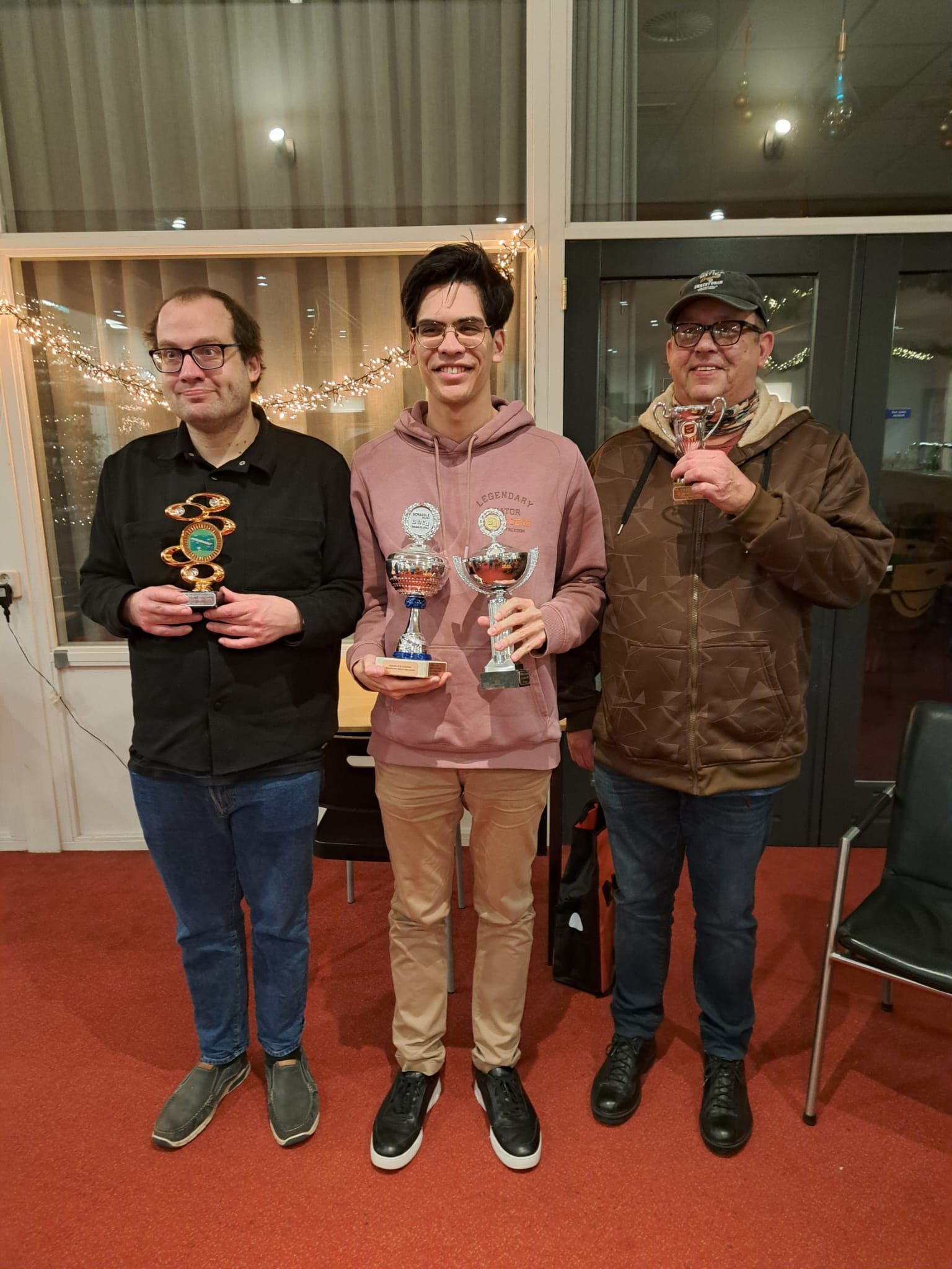 Drie mensen met trofeeën staan ​​op een rode loper. De middelste is jong, de anderen zijn ouder. Goed verlichte ruimte.
