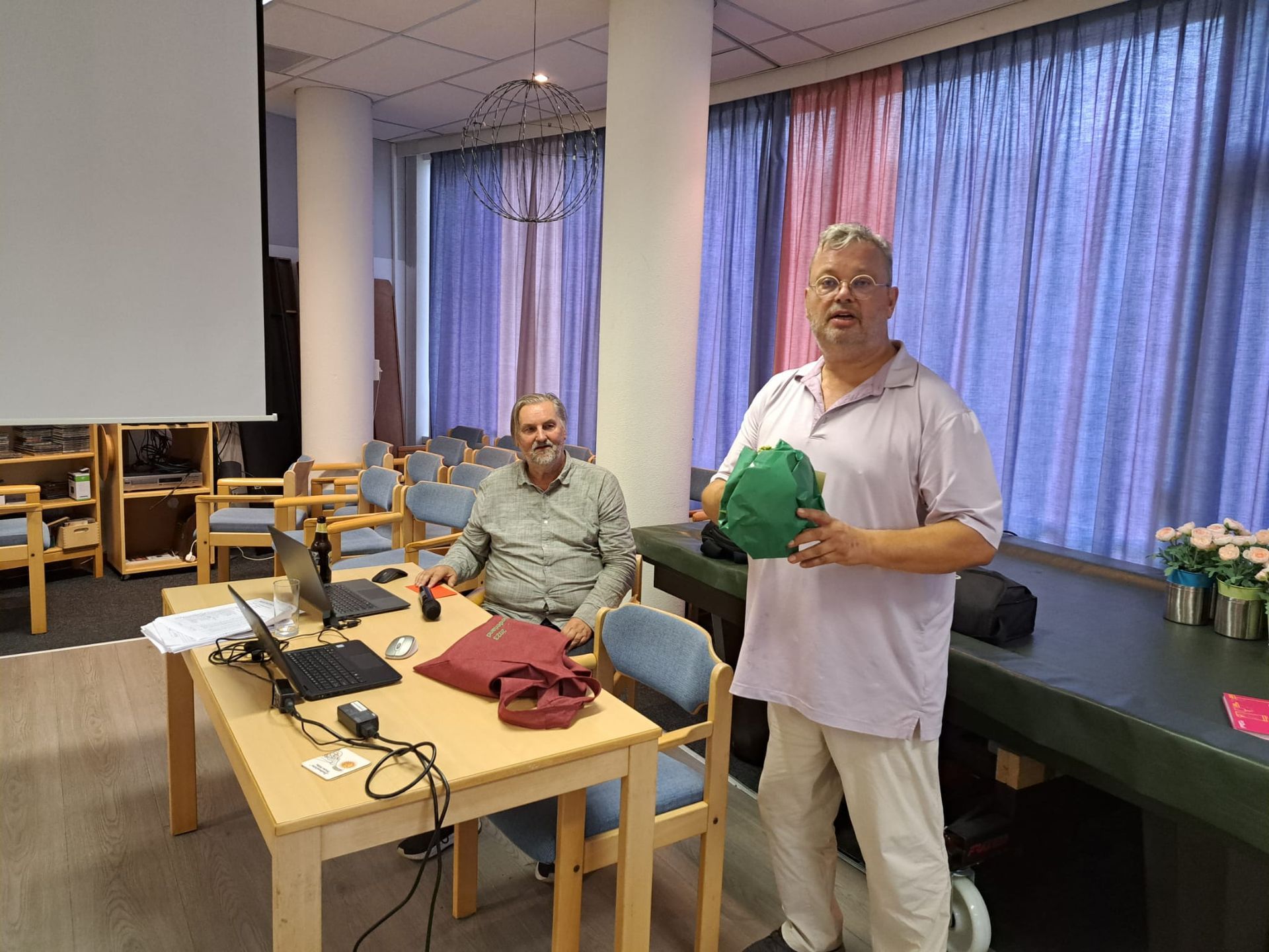 Twee mannen zitten aan een tafel met laptops, een van hen houdt een groen voorwerp vast, binnen met gordijnen en een projectiescherm.