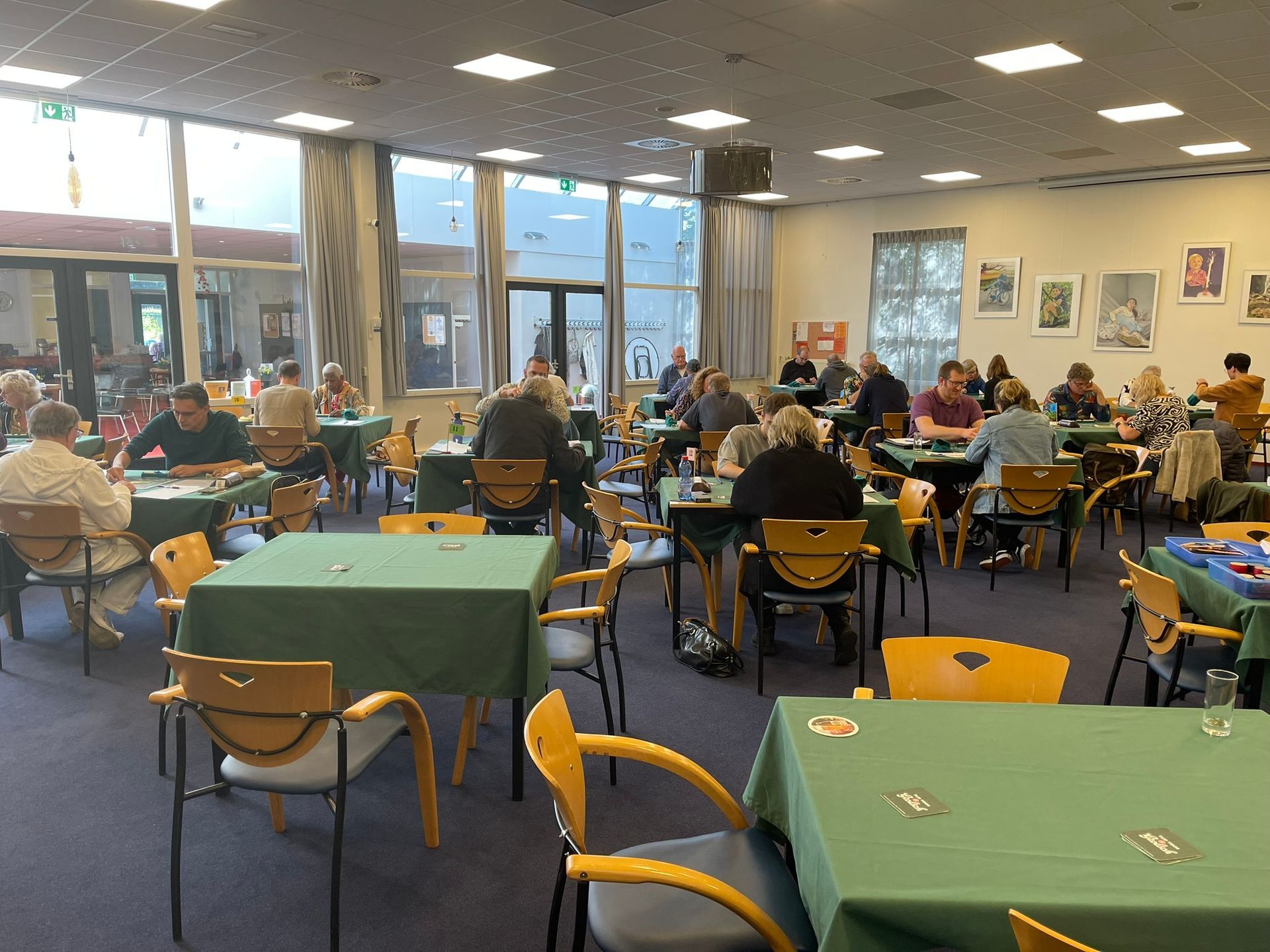 Mensen zitten aan tafels in een felverlichte ruimte en spelen spelletjes. Groene tafelkleden, kunst aan de muren en grote ramen.