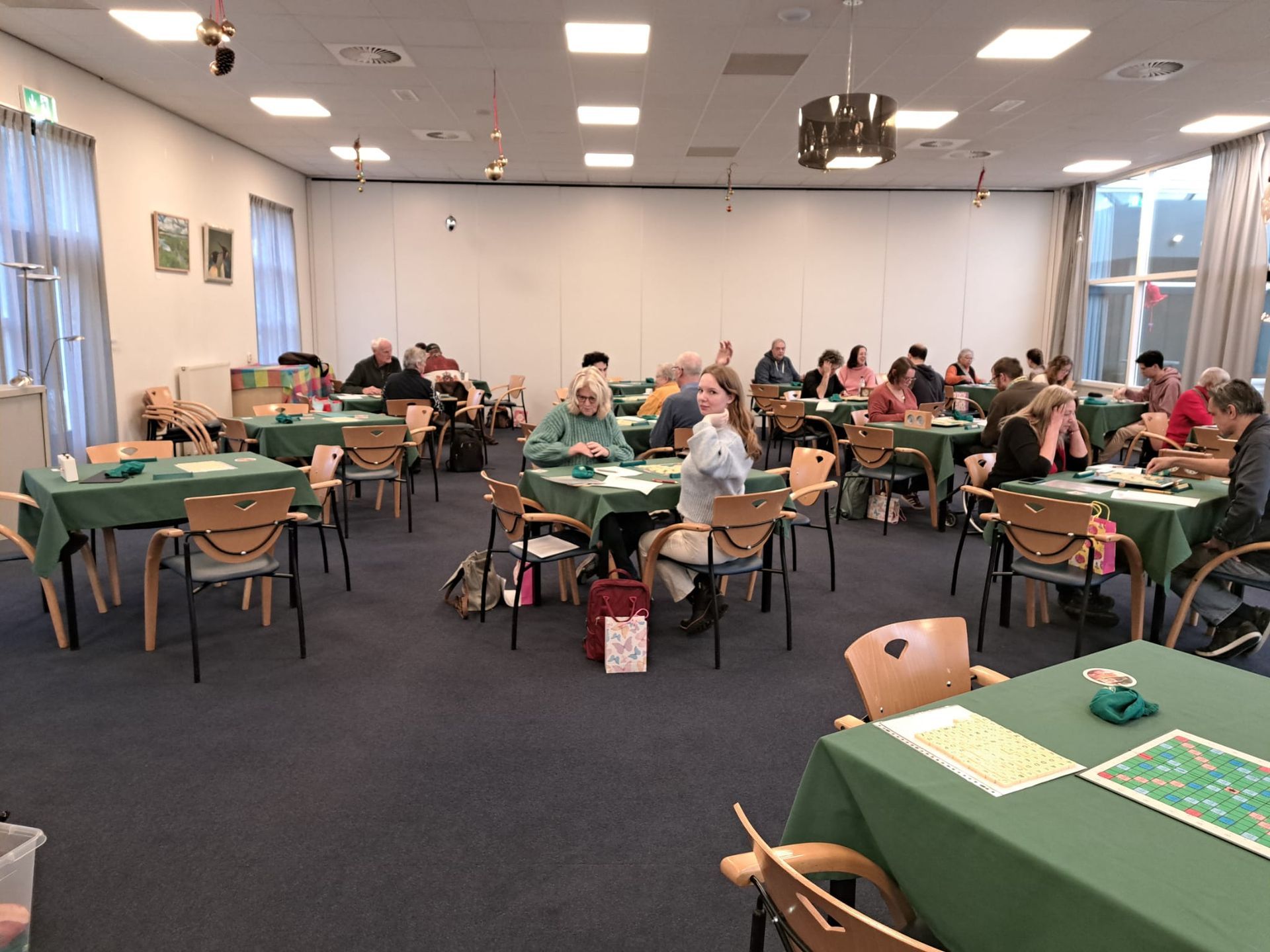 Mensen zitten aan tafels en spelen een spel in een goed verlichte ruimte. Groene tafelkleden, neutrale muren, blauw tapijt.