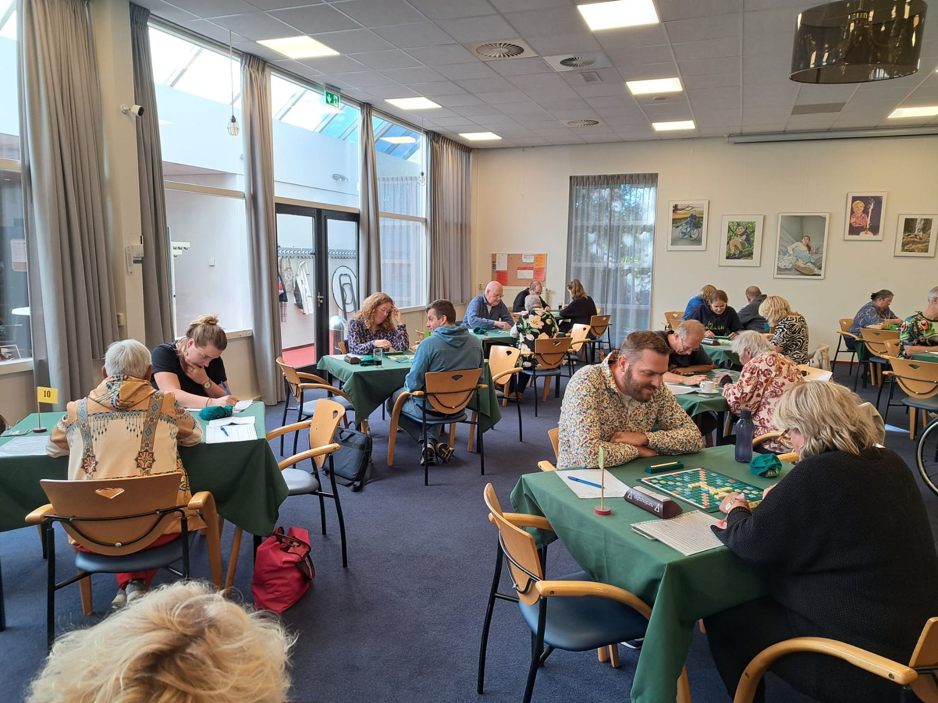 Mensen spelen bordspellen aan tafels in een felverlichte ruimte. Groene tafelkleden, verschillende gezichtsuitdrukkingen en geconcentreerde bewegingen zijn duidelijk zichtbaar.